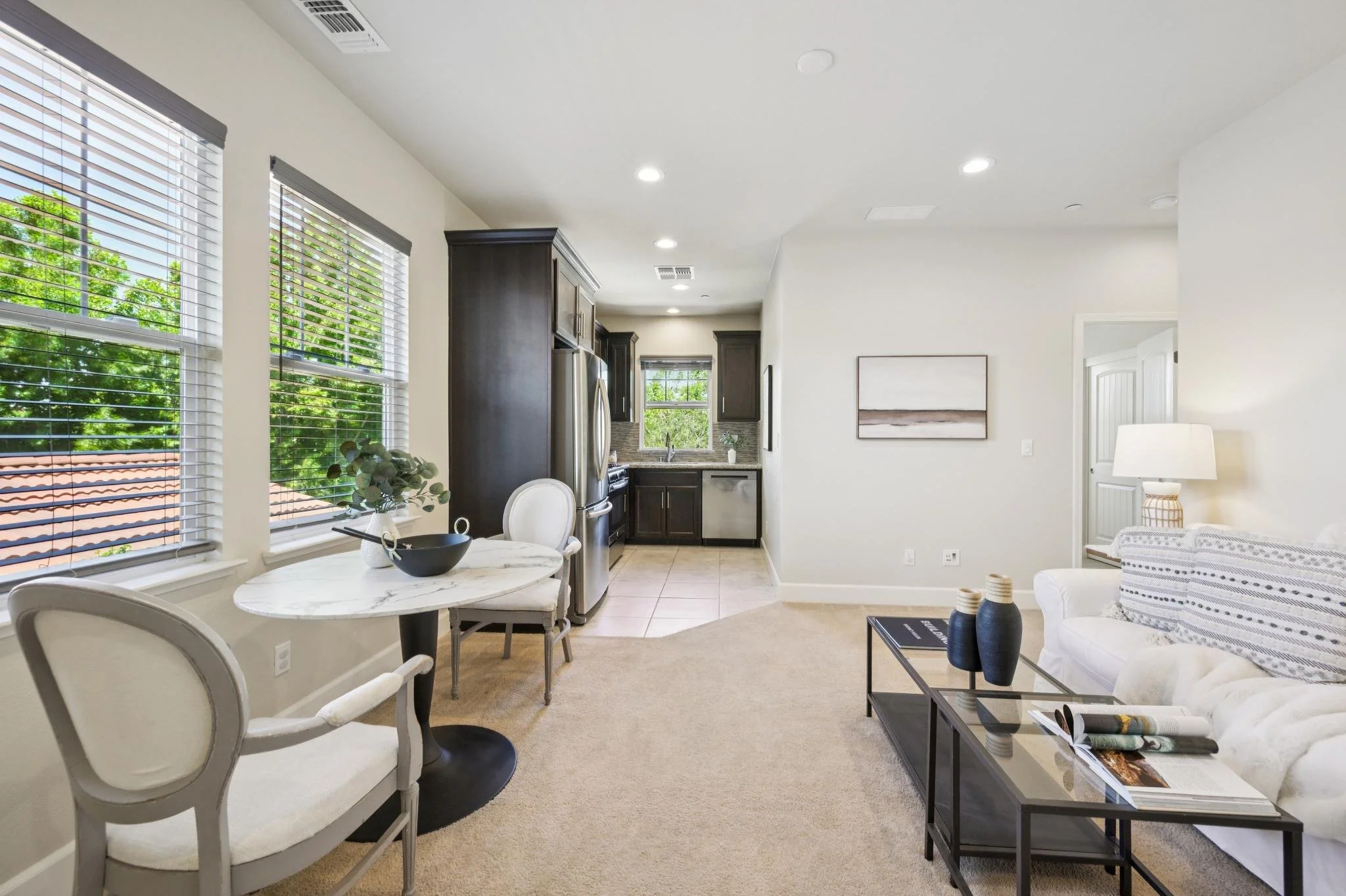 Open-concept living space with kitchen and dining areas, featuring large windows with blinds, dark cabinets in the kitchen, white walls, and a mix of furniture including a round marble-top dining table with white chairs, a white sofa with striped cushions, and a black coffee table with decorative vases and magazines.