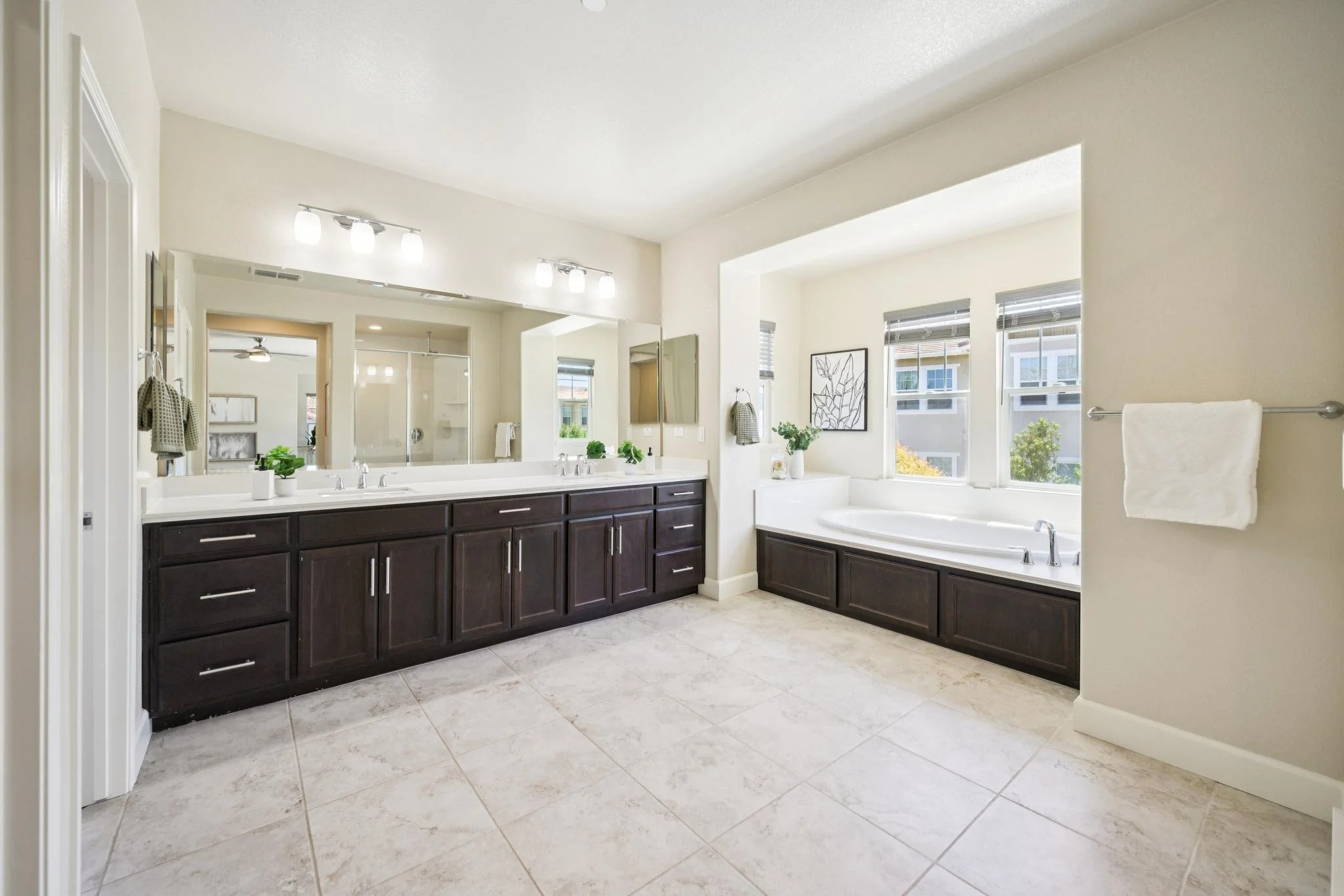 Modern bathroom with dark wood vanity, large mirror, double sinks, white countertop, bathtub with windows, and neutral color scheme.