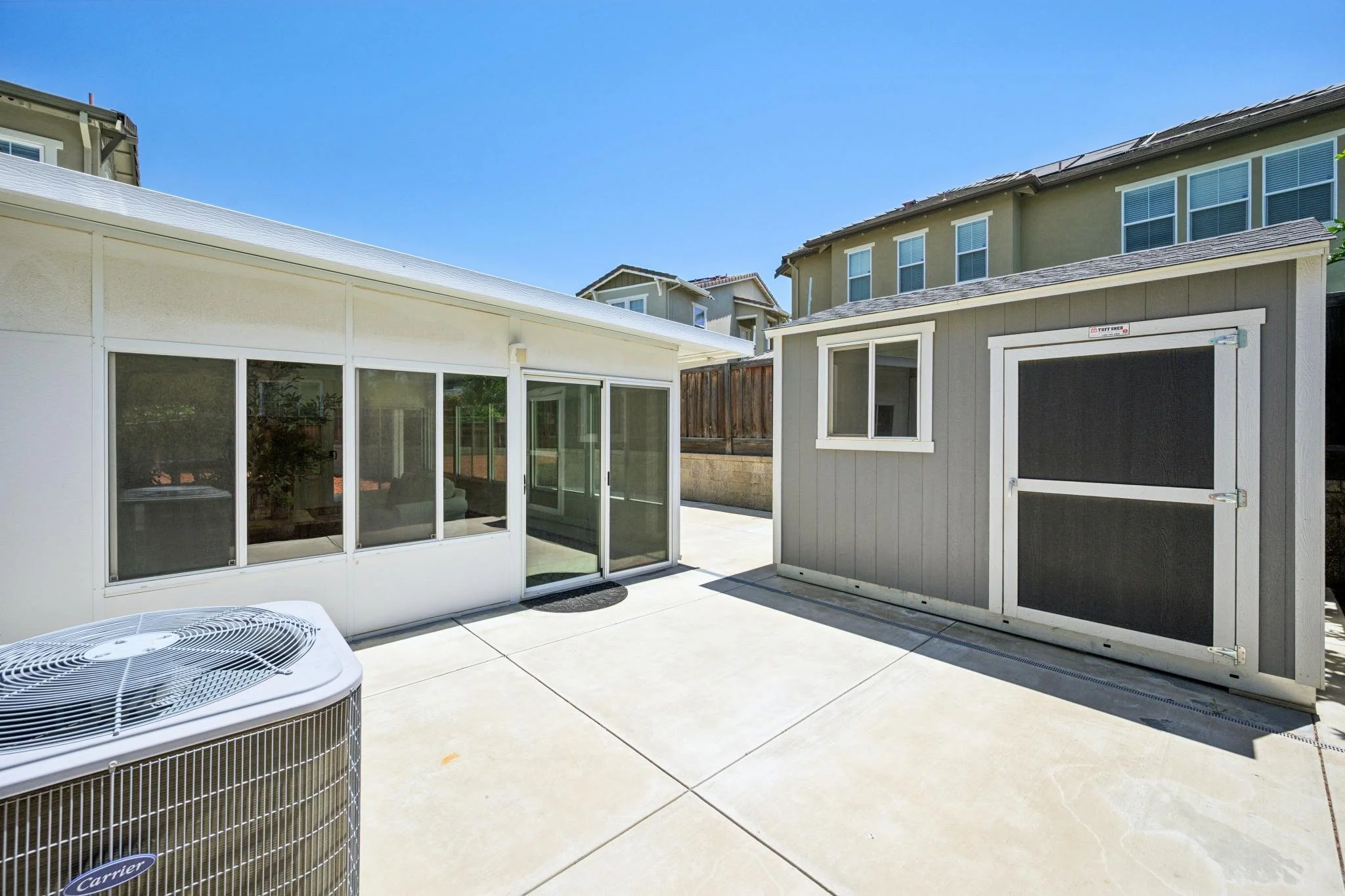 View of a backyard patio area with two small storage sheds, one white with glass windows and a door, and one gray with a sliding window. An air conditioning unit is visible in the foreground, and there are multiple residential houses in the backgroun
