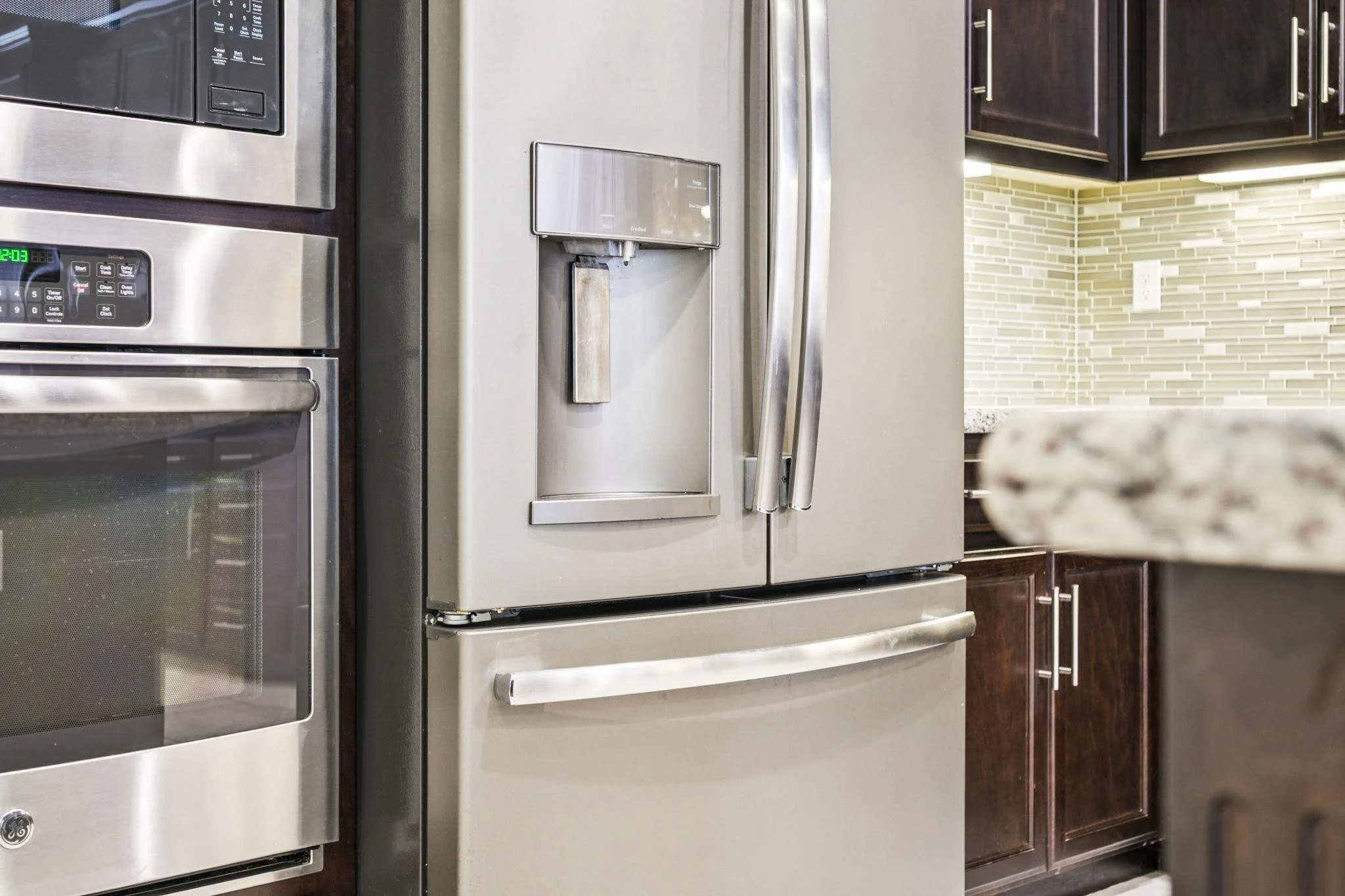 A stainless steel side-by-side refrigerator in a kitchen with dark wood cabinets and a beige tile backsplash.