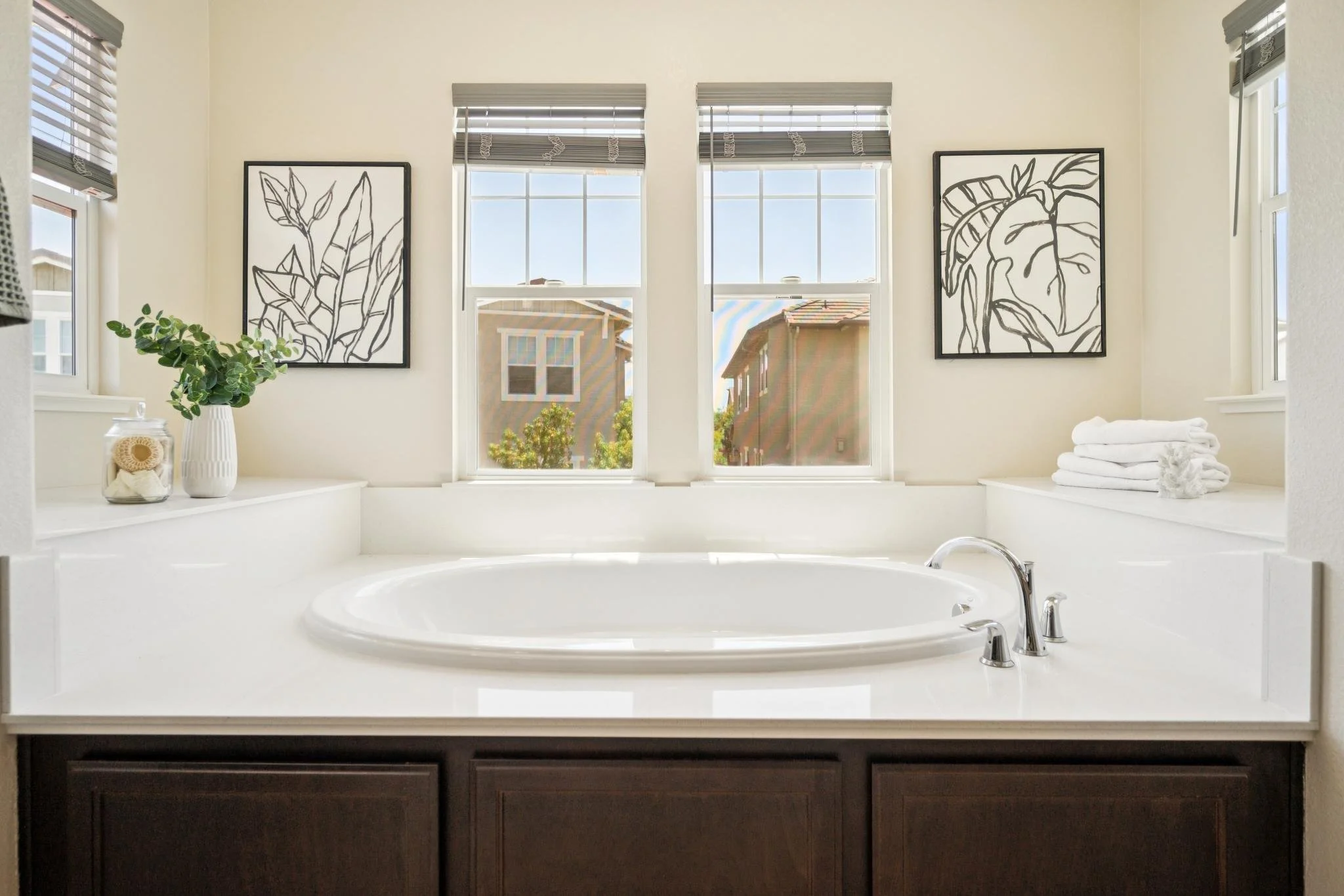 A bathroom with a large oval bathtub set into a white surround, two windows with gray blinds, framed artwork of black line drawings of leaves, a white vase with green foliage, a jar with bath supplies, and stacked white towels.