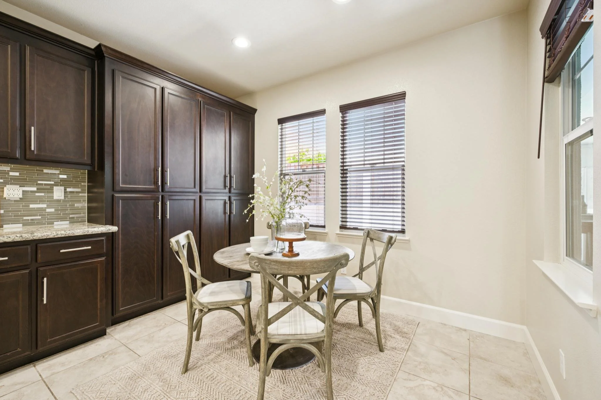 A kitchen corner with dark wood cabinets, a beige tile floor, a round wooden dining table with four chairs, and windows with brown blinds.