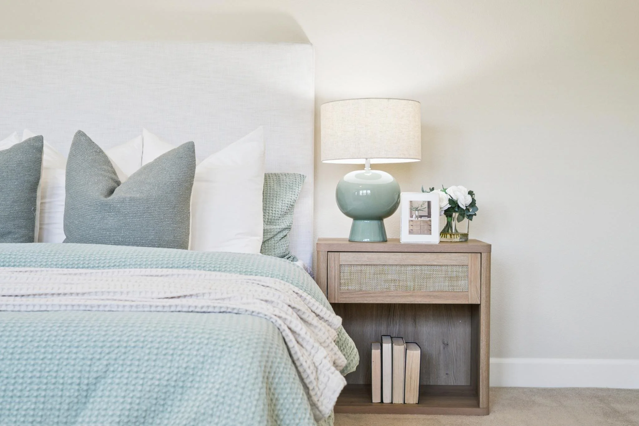 A bedroom with a bed featuring gray and white pillows, a wooden nightstand with books, a lamp, a framed photo, and white flowers, against a light-colored wall.