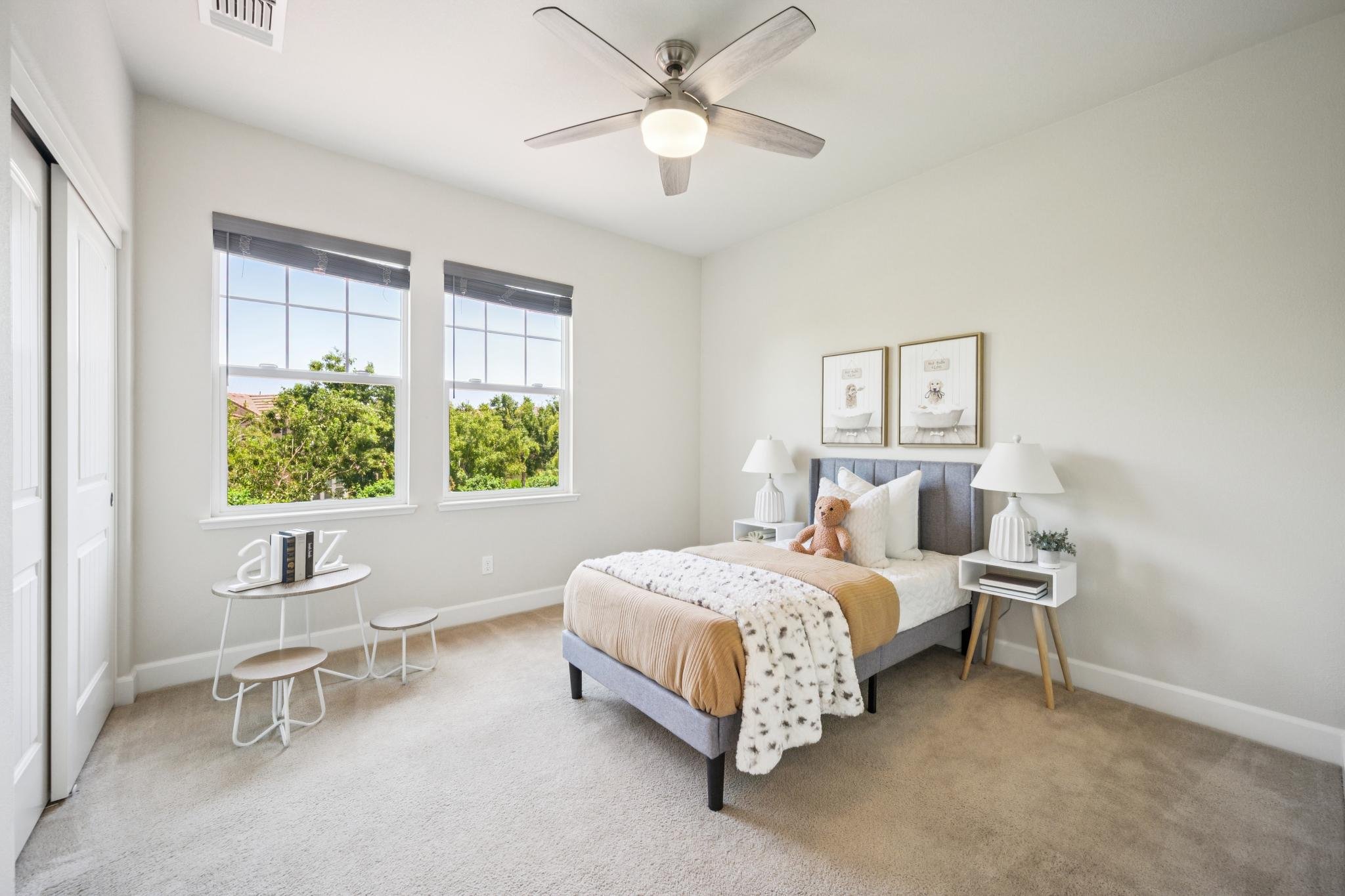 Cozy bedroom with a bed, nightstand, lamps, and wall art, bright windows showing trees outside, ceiling fan, and neutral decor.