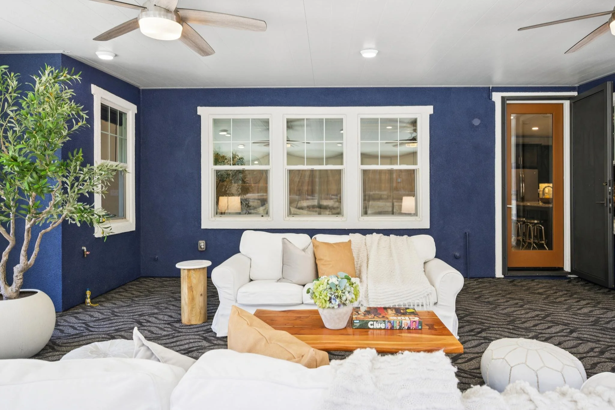 A cozy living room with navy blue walls and three white-framed windows. There's a white sofa with beige and white pillows, a wooden coffee table with a floral arrangement and a Clue board game, and a large potted plant. The room has a patterned carpe