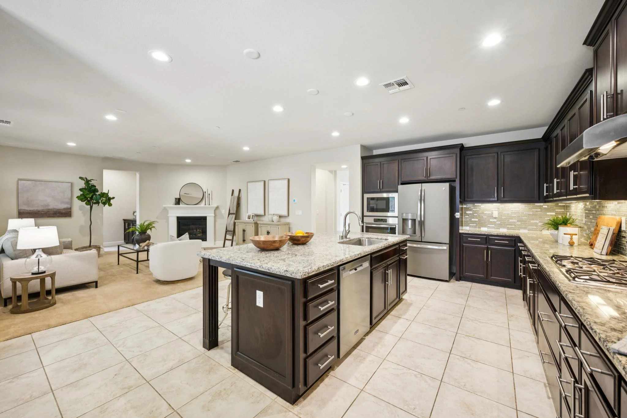 Modern kitchen with dark wood cabinets, granite countertops, and stainless steel appliances, connected to a living area with a fireplace and beige seating.