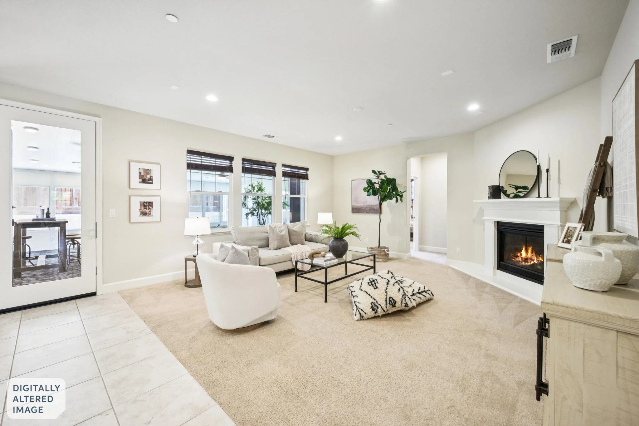 Bright living room with white walls, three windows with dark blinds, a white sofa with gray cushions, a black coffee table with books and a plant, a white armchair, a patterned rug, and a fireplace with a mirror above it. Decor includes framed pictur
