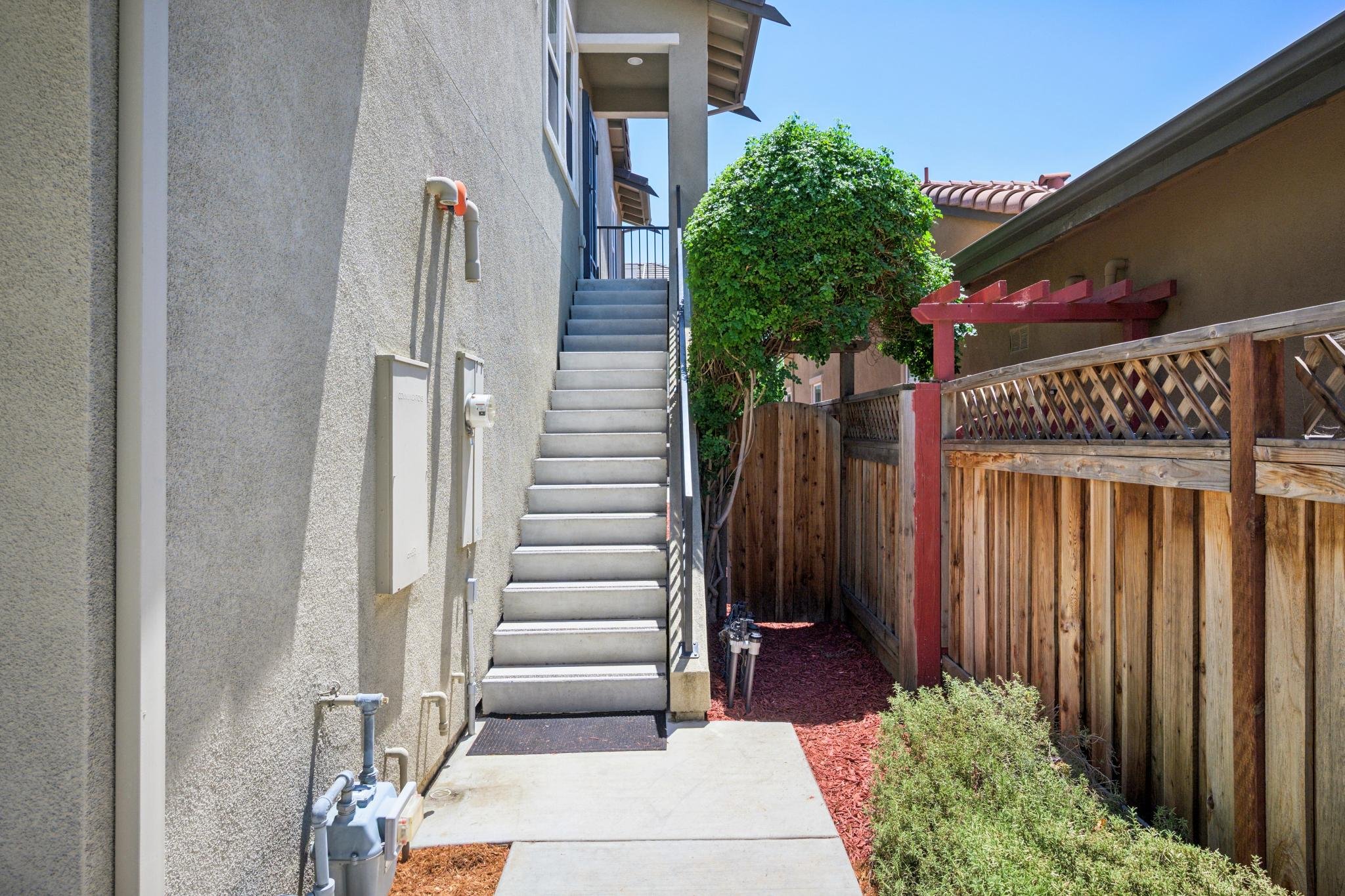 Backyard side view showing a light grey house wall with a staircase leading to a second floor, a wooden fence with a lattice top, a lush green tree, and a clear blue sky.