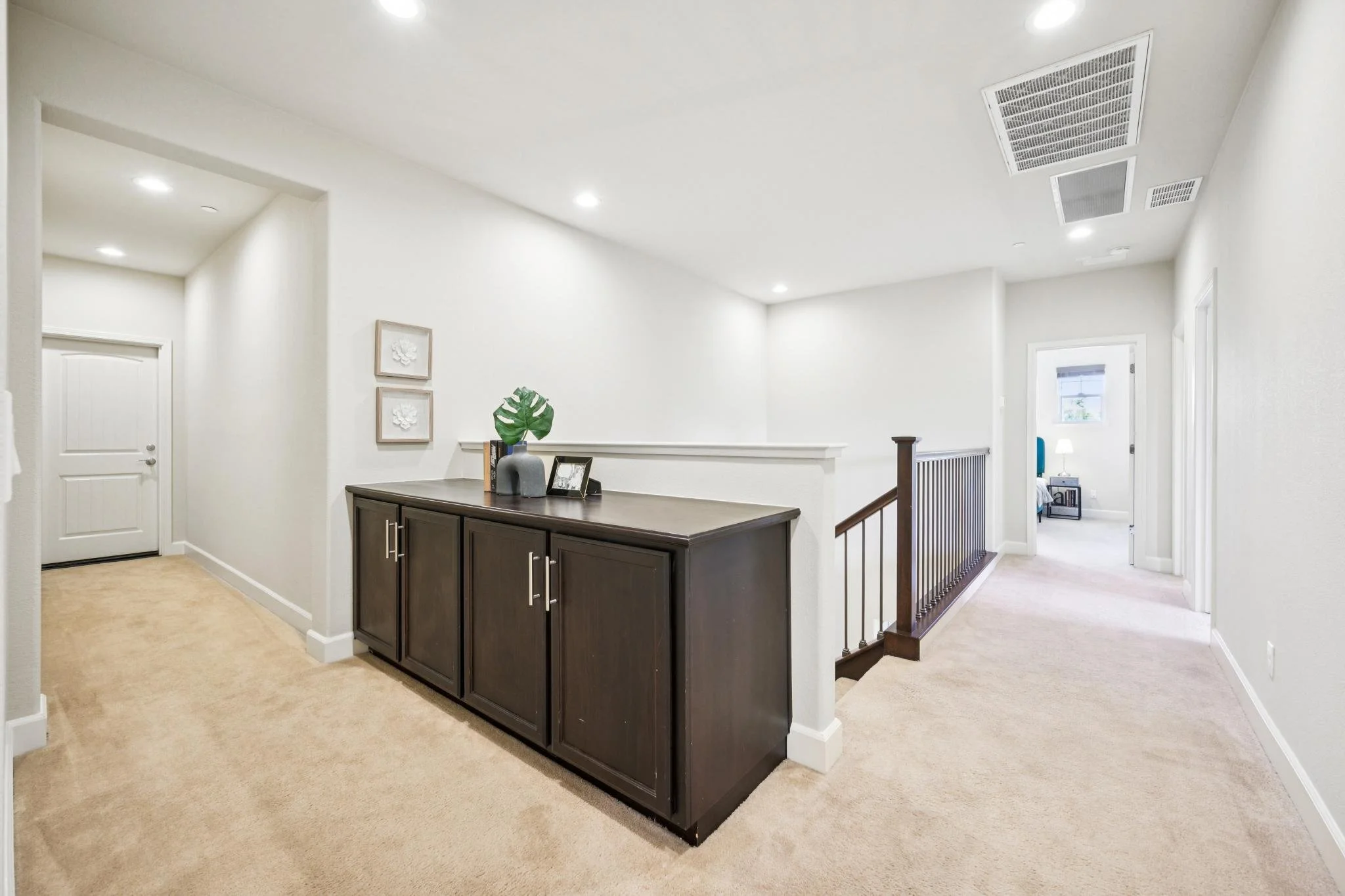 Interior view of a hallway with a decorative dark wood sideboard, featuring a vase with large green leaves, a stack of books, and picture frames. The space is brightly lit with multiple ceiling lights, white walls, plush beige carpet, and an open doo
