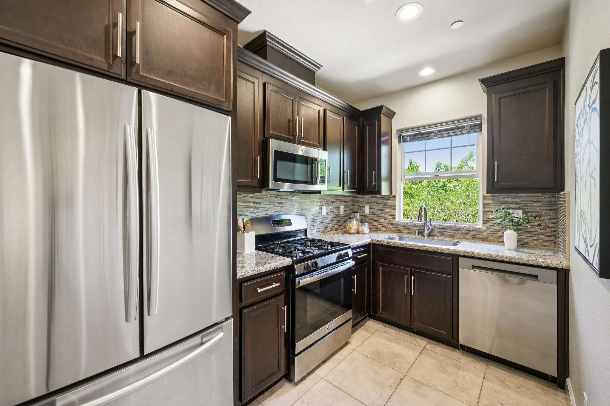 A modern kitchen with dark wood cabinets, a stainless steel microwave, and a gas stove. A window with a view of trees, granite countertops, and a small white potted plant are visible.