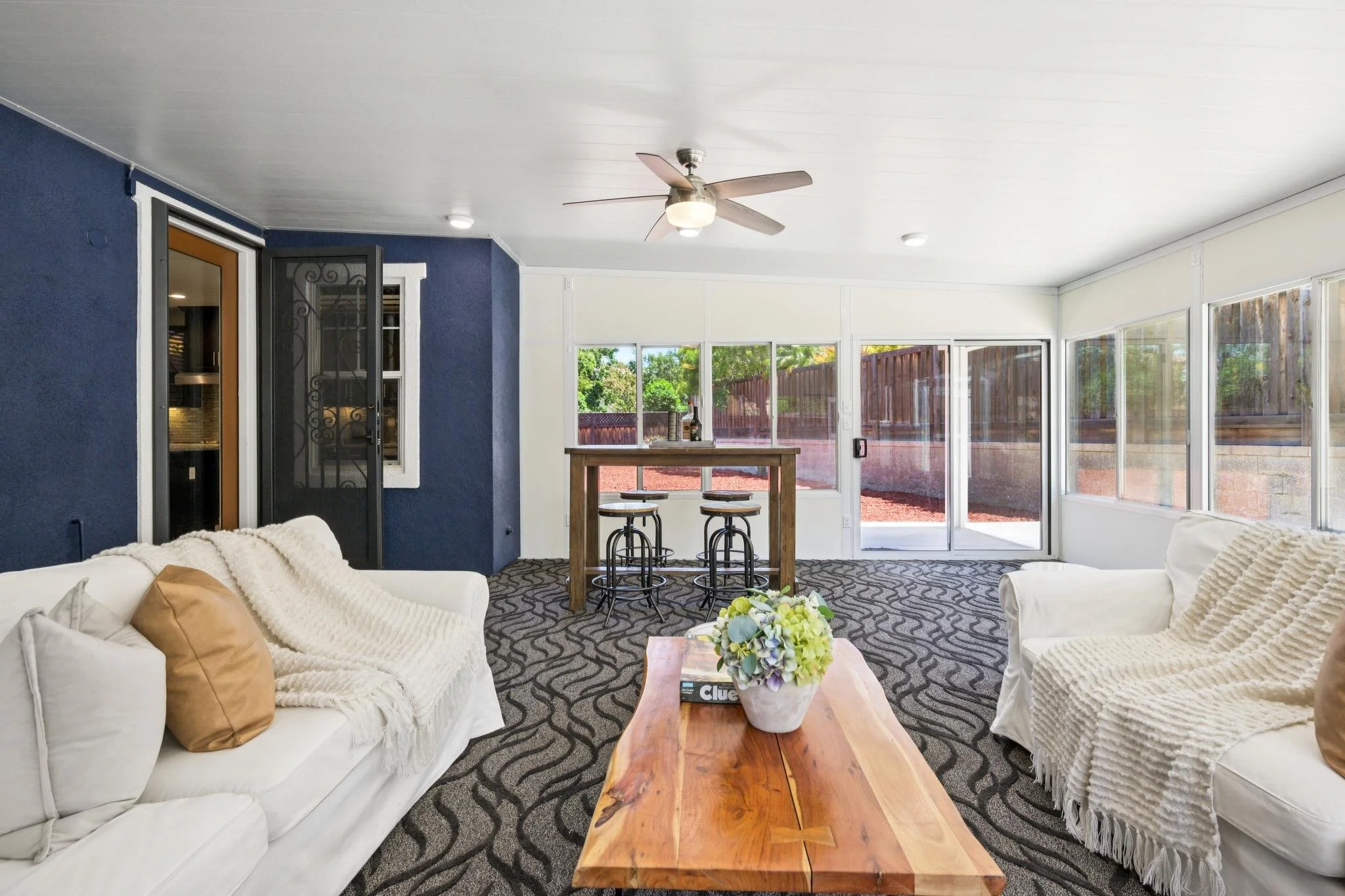 Living room with white sofas, beige pillows, a wooden coffee table with a floral arrangement, patterned carpet, blue and white walls, ceiling fan, large windows, and a door leading outside.