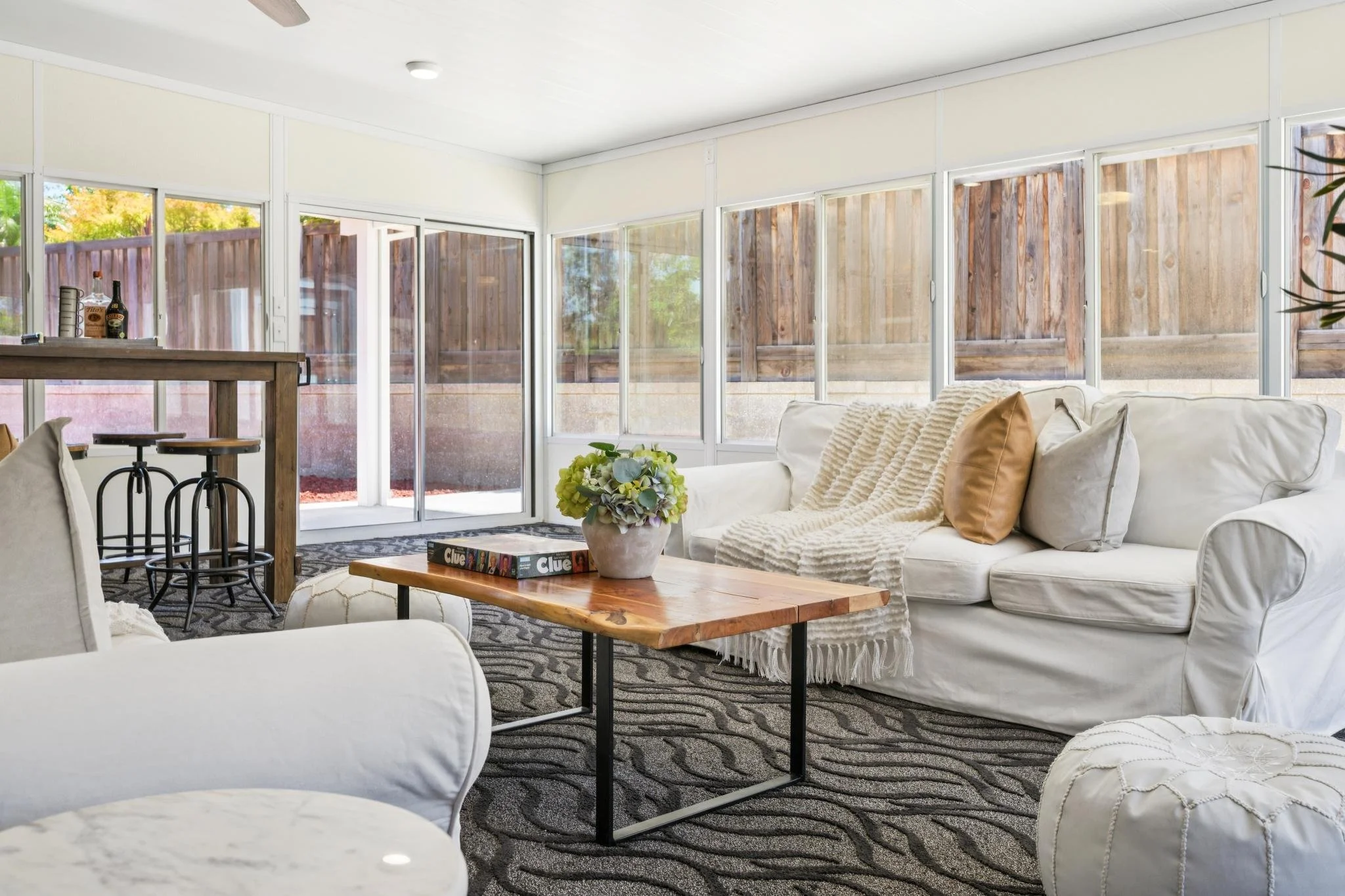 Bright living room with large windows showing wooden privacy fence, white sofa with beige pillows, wooden coffee table with a flower vase, patterned rug, and bar stools near a wooden counter.