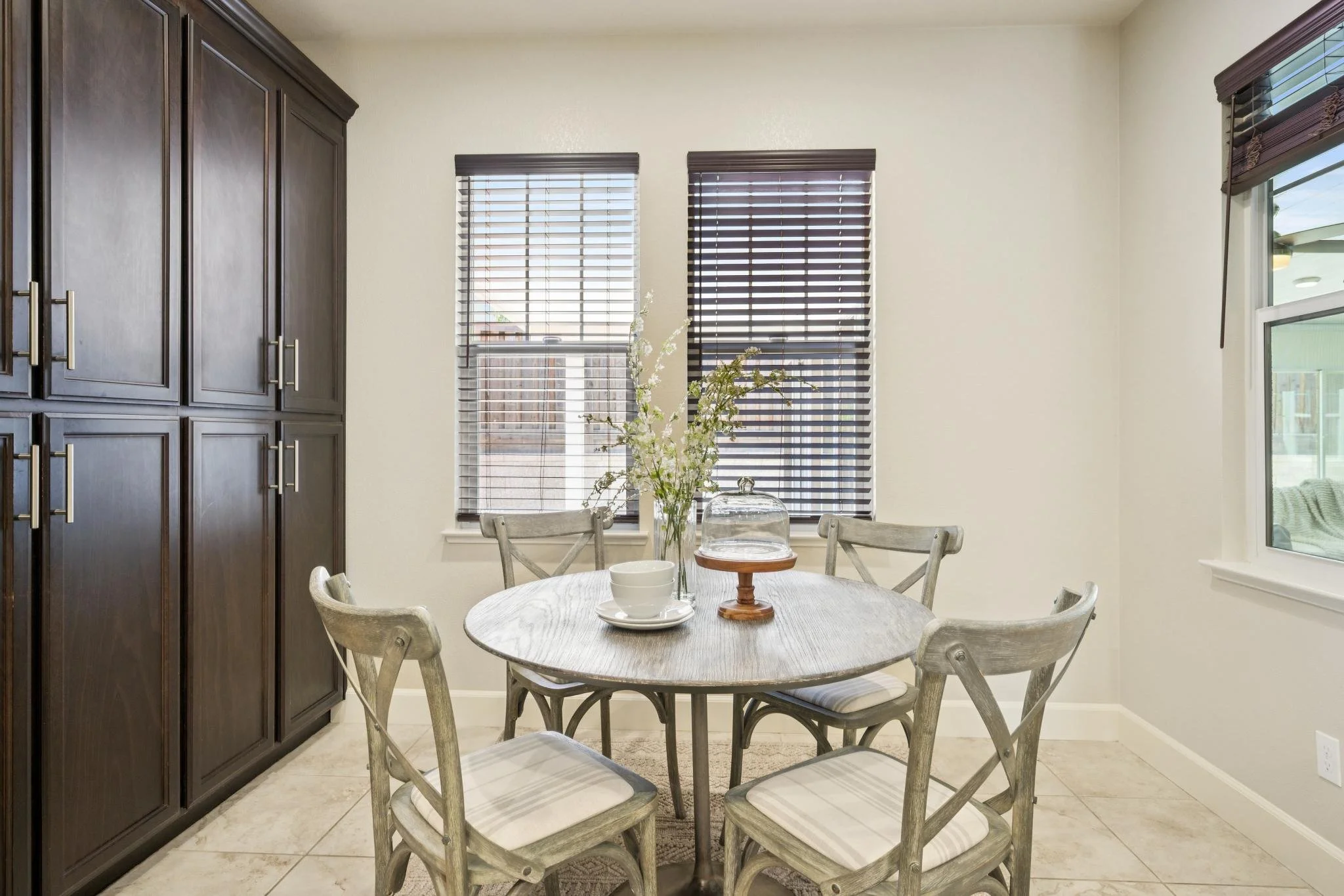 A dining area with a round wooden table, six matching chairs with white cushions, a glass vase with white flowers on the table, and dark wooden cabinetry to the left. There are three windows with blinds, and the walls are cream-colored with tiled flo