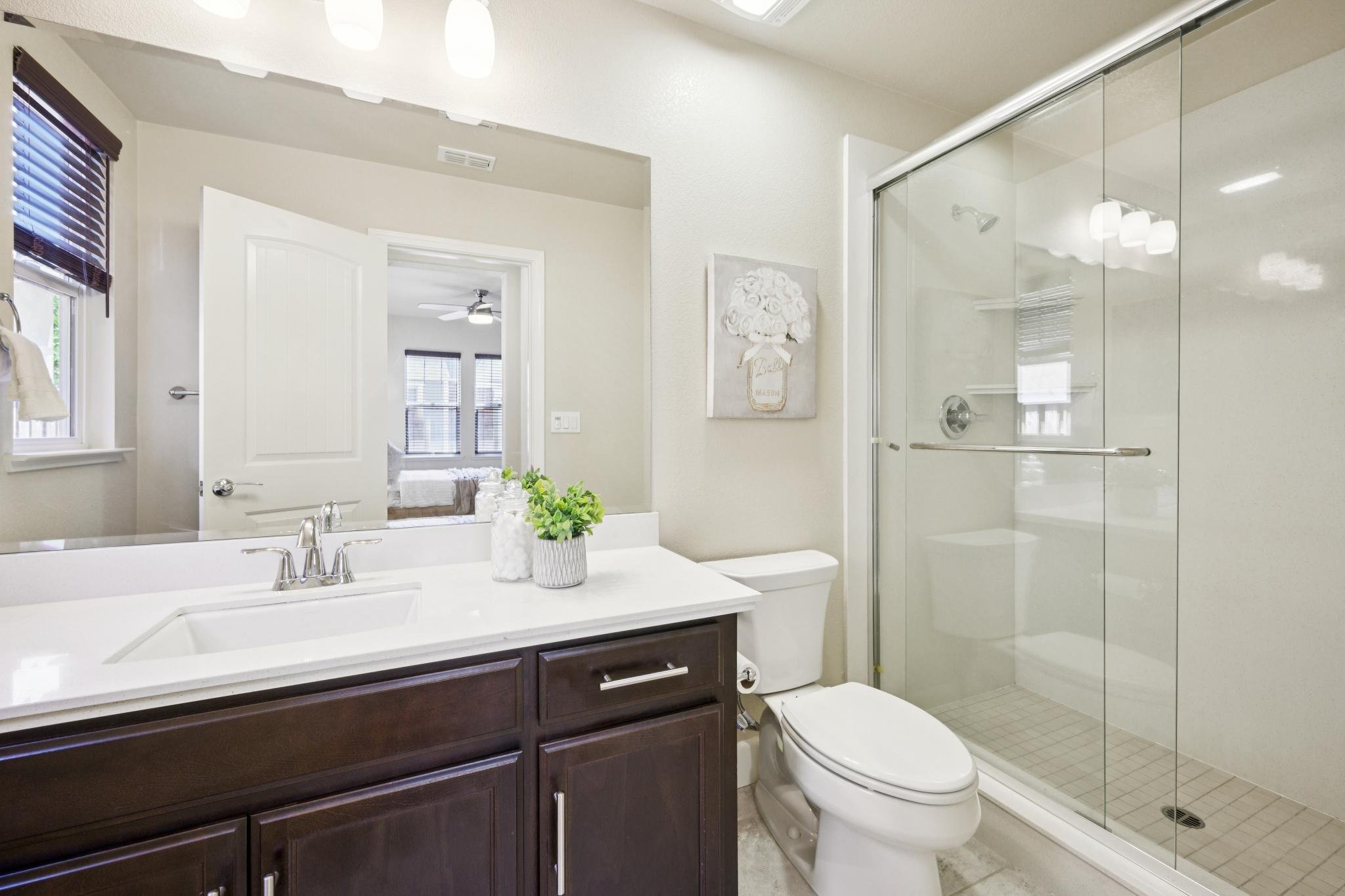 A modern bathroom with a dark wooden vanity, white countertop, and a large mirror. A white toilet is beside the sink. There is a glass-enclosed shower with a door on the right. Two small potted plants on the countertop, and framed art on the wall. Wi
