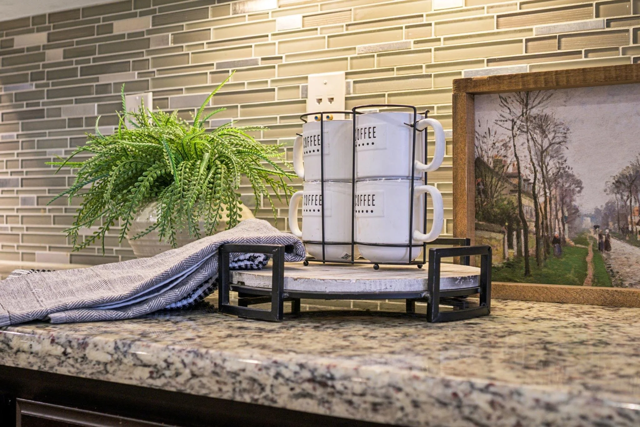 Decorative kitchen countertop with a potted green plant, a set of four white coffee mugs with black text on a black metal mug rack, a cloth, and a framed landscape painting of trees and a pathway against a tiled wall.