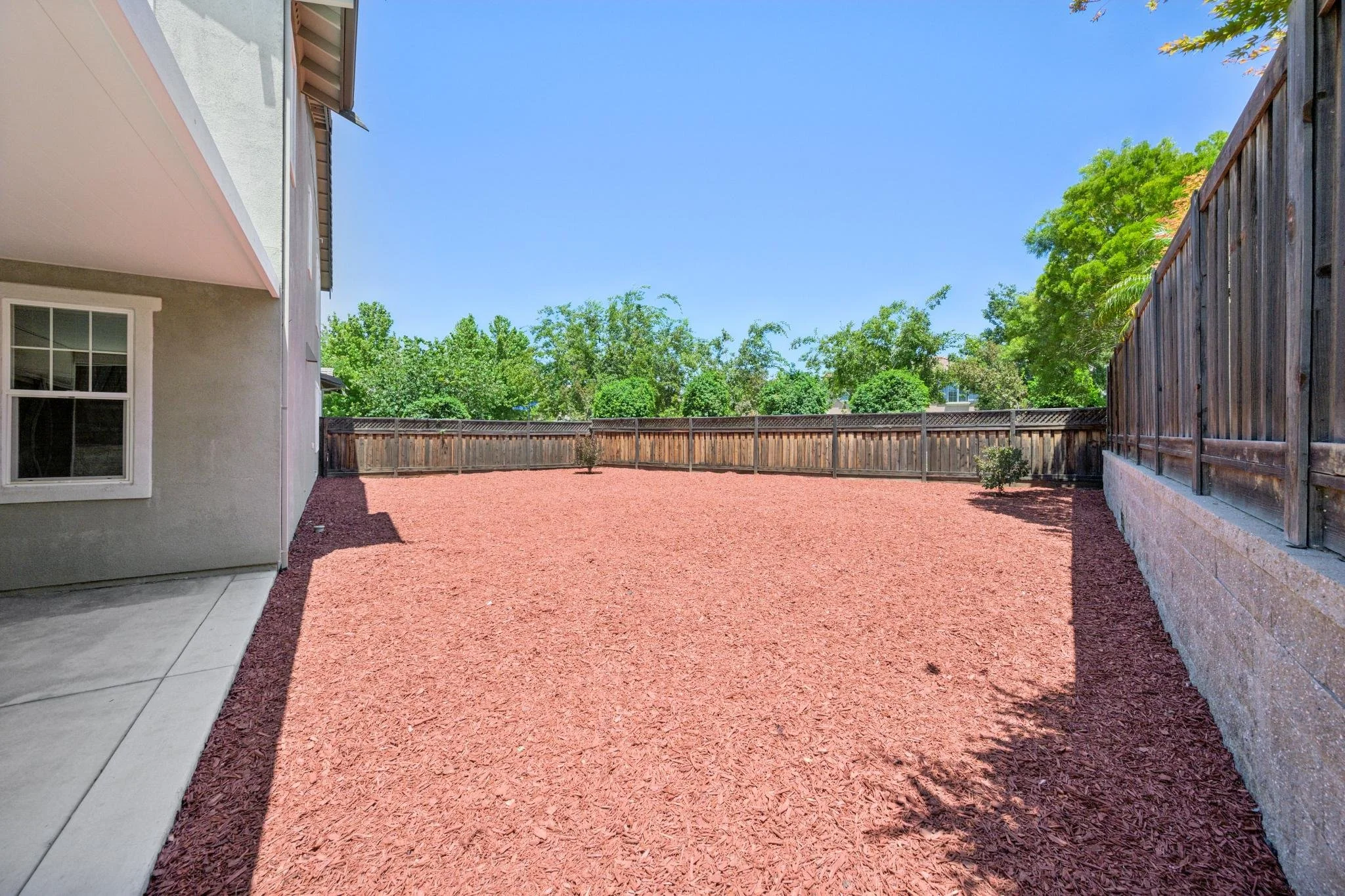 A backyard with a reddish-brown gravel yard, a wooden fence, and trees in the background. Part of a house with a window and a concrete patio on the left side.