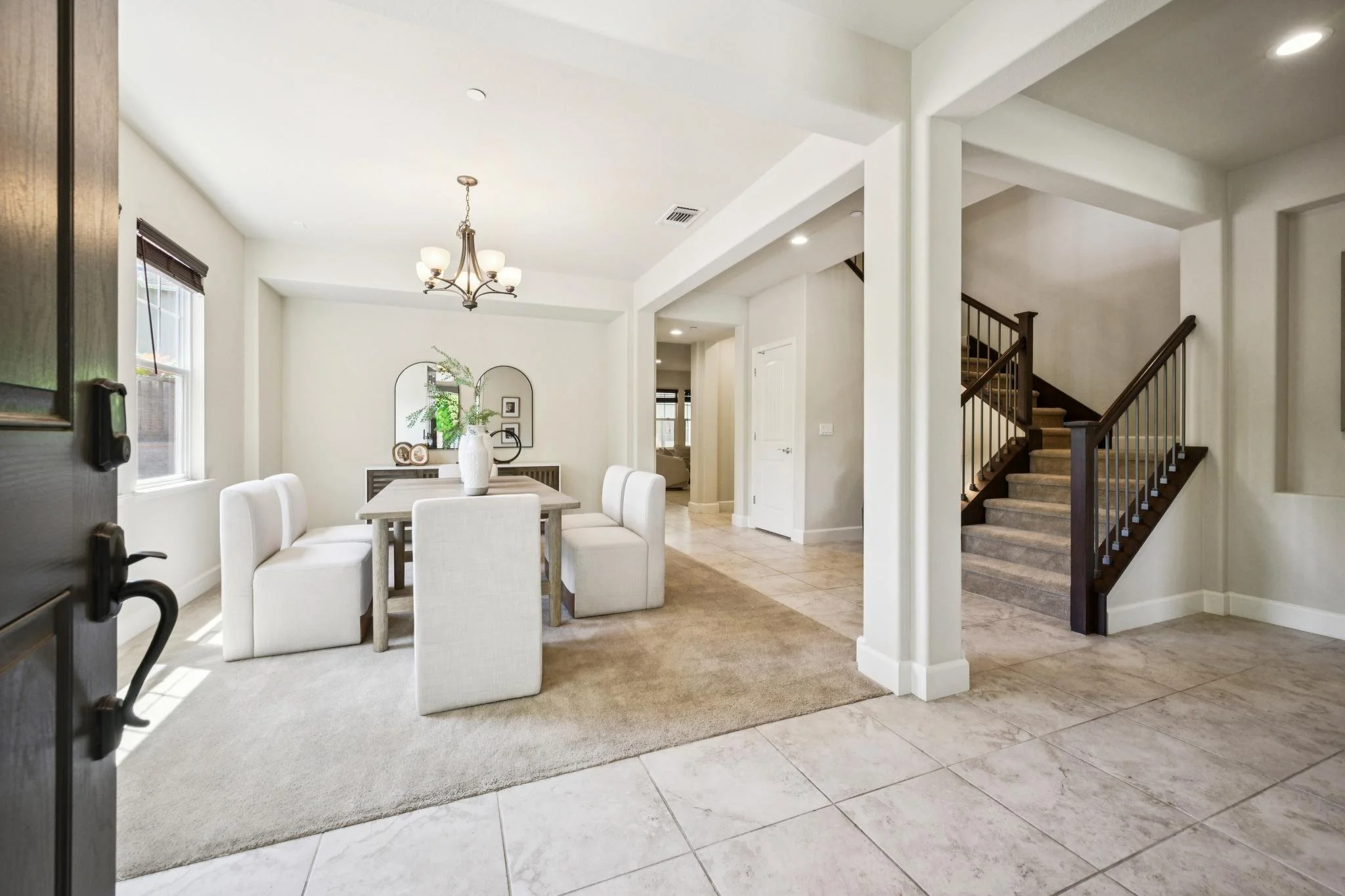 A view of a modern, bright living space with a white dining table and chairs, decorative vases, wall mirrors, staircase, and natural light coming through windows.