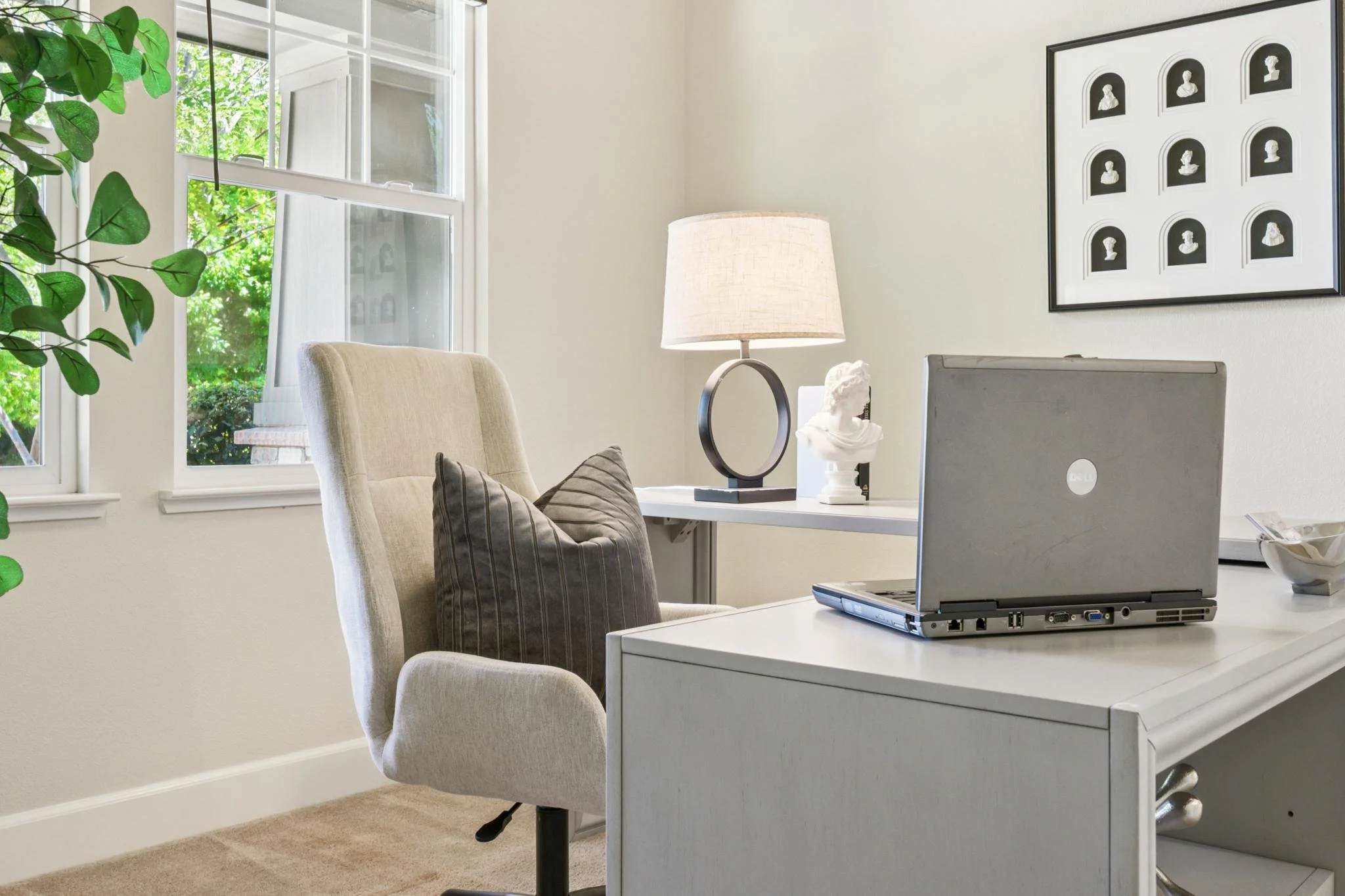 A home office with a white desk holding a silver laptop, a white bust sculpture, a black and white table lamp, and a glass bowl. Behind the desk is a beige upholstered chair with striped pillows. The wall features a framed black-and-white collage of 
