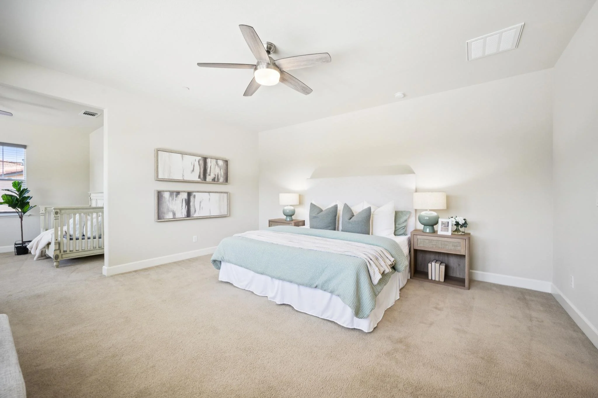 Bedroom featuring a bed with a light blue bedspread, two bedside tables with lamps, and wall art. An adjoining area with a crib and a window with blinds is visible.