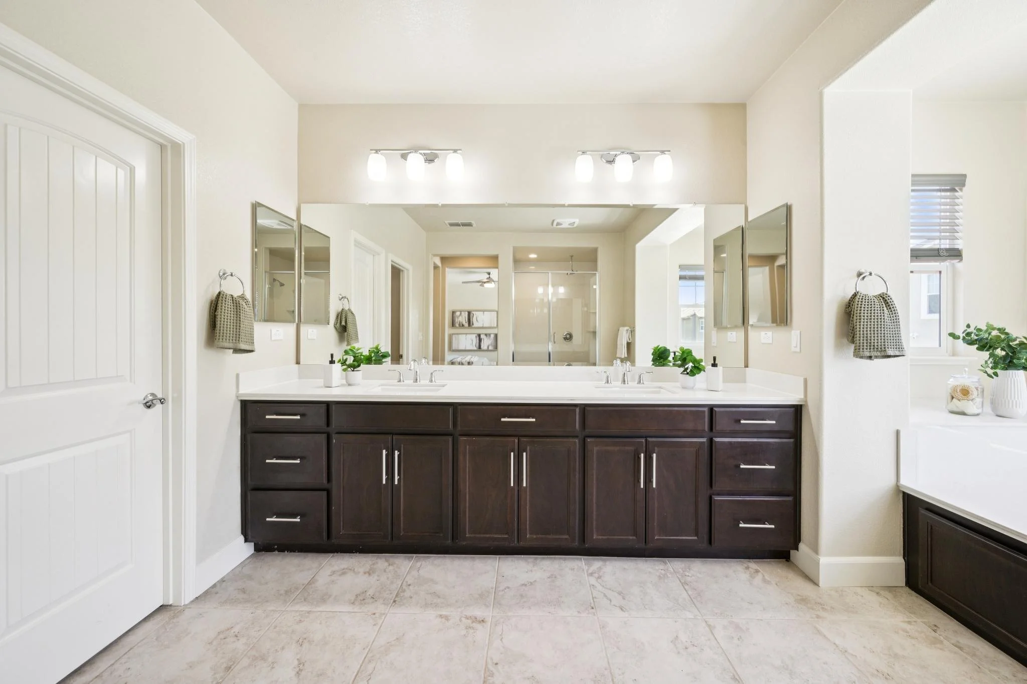 Bathroom with double vanity, large mirror, dark wood cabinets, and two wall-mounted light fixtures. Walls are painted light, with tiled floor and small plants on the counter.