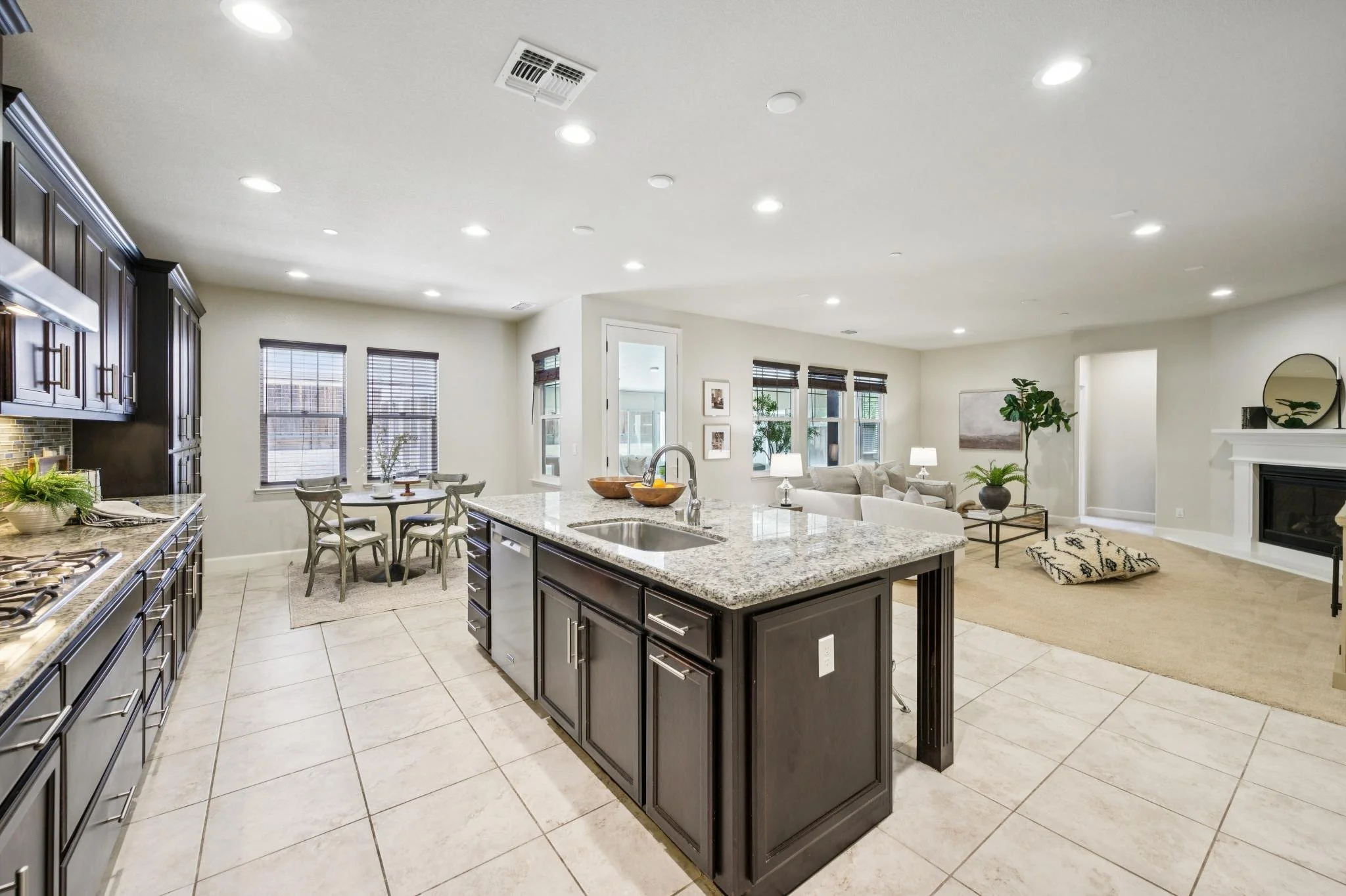 Open-concept kitchen and living room with dark cabinets, granite countertops, dining area, and beige carpet with seating area near white fireplace and windows.