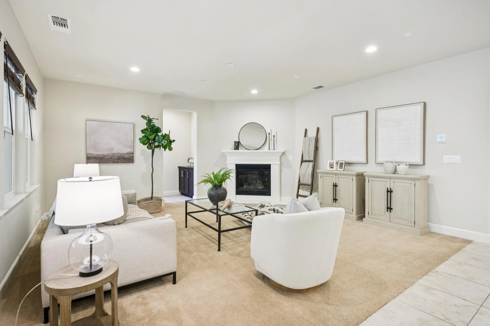 A spacious living room with white walls, beige carpet, and modern furniture including white armchairs, a glass coffee table, and side tables with lamps. Decor includes framed artwork, potted plants, a fireplace, and wooden furniture pieces.