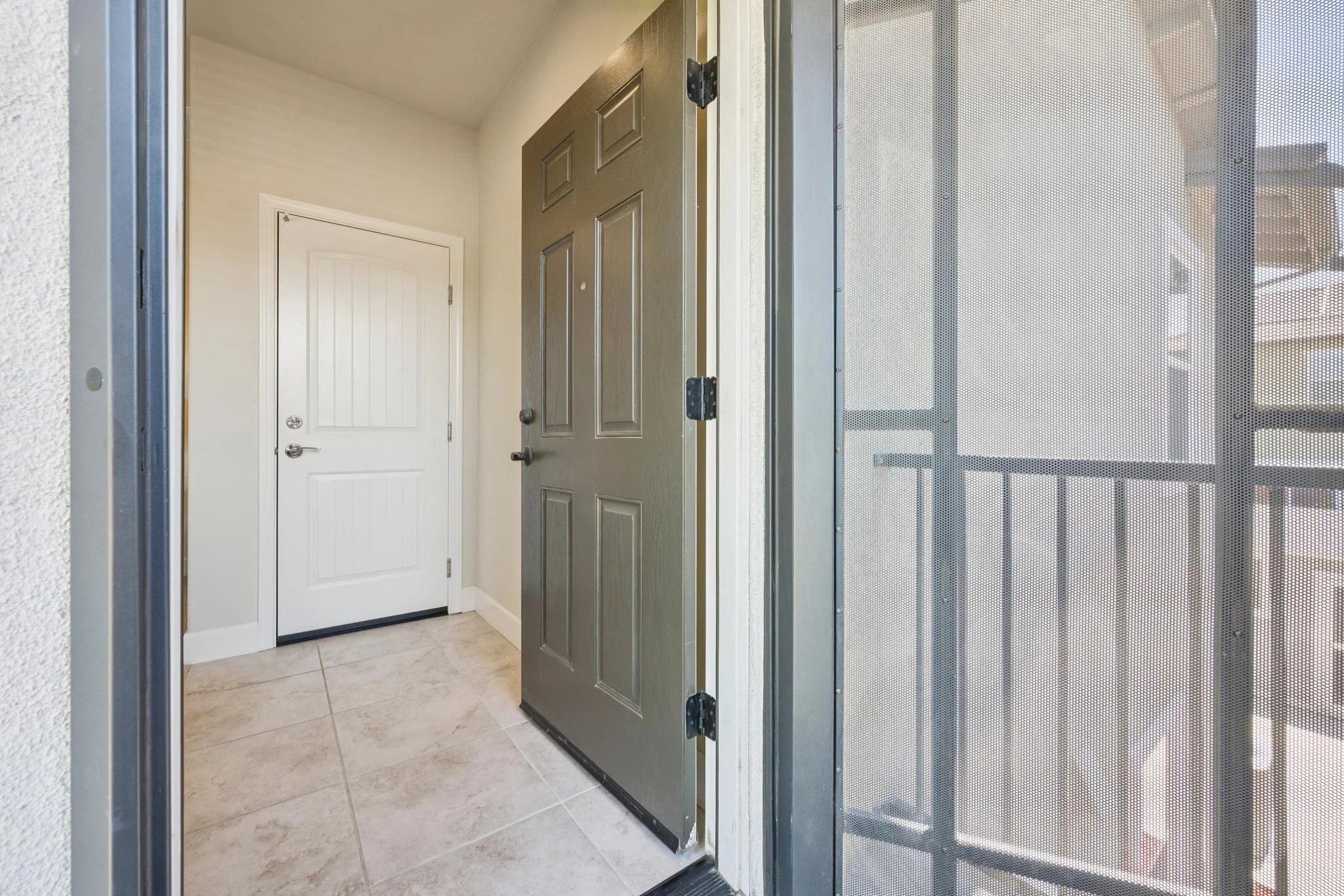 View of an apartment or condo entrance from the inside, showing a brown door and a white door, with a screen door on the right side. The floor is tiled and the walls are light-colored.