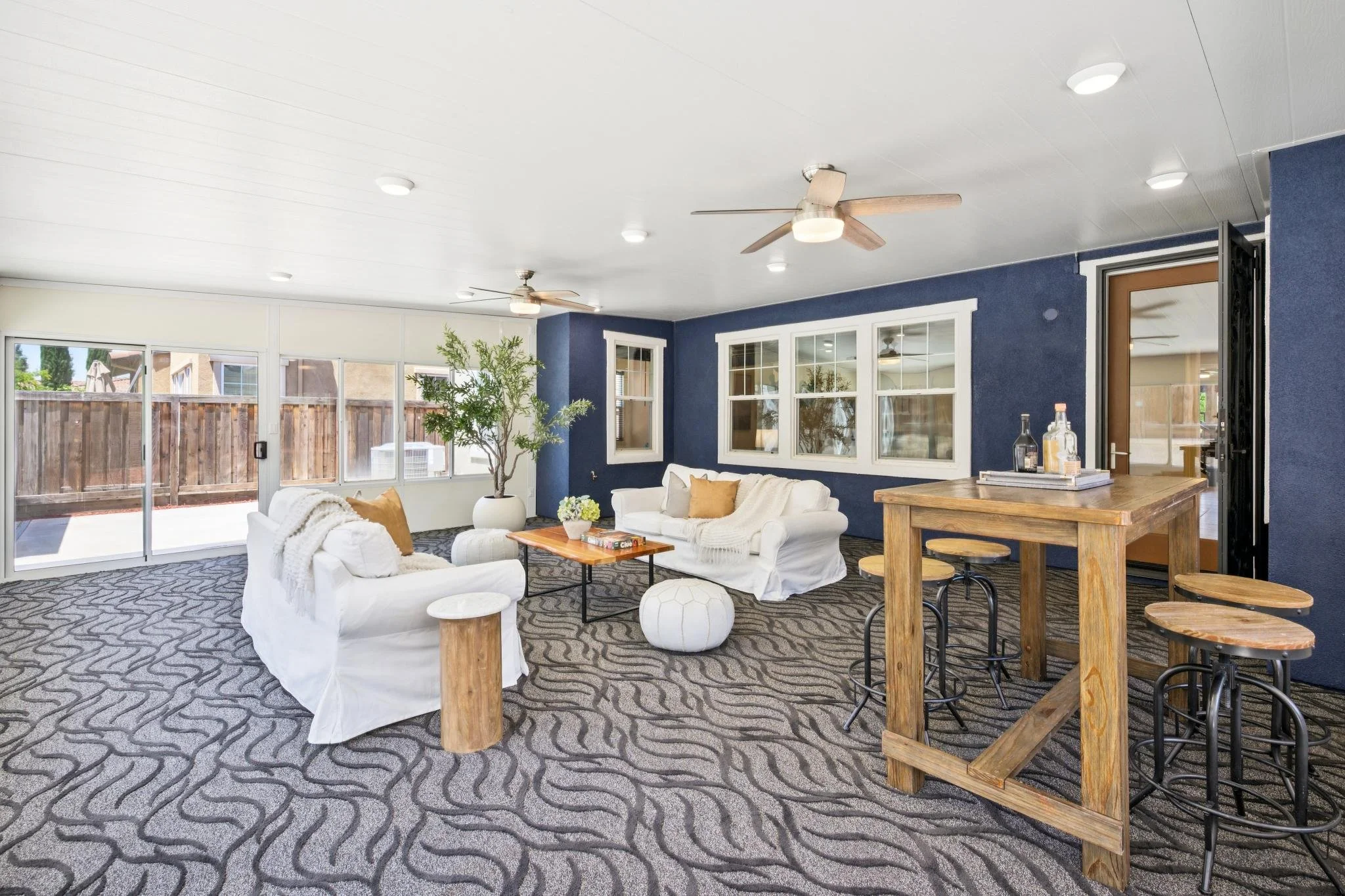Living room with white sofas, throw pillows, a coffee table, a potted plant, large windows, a sliding glass door, ceiling fans, and a patterned carpet.