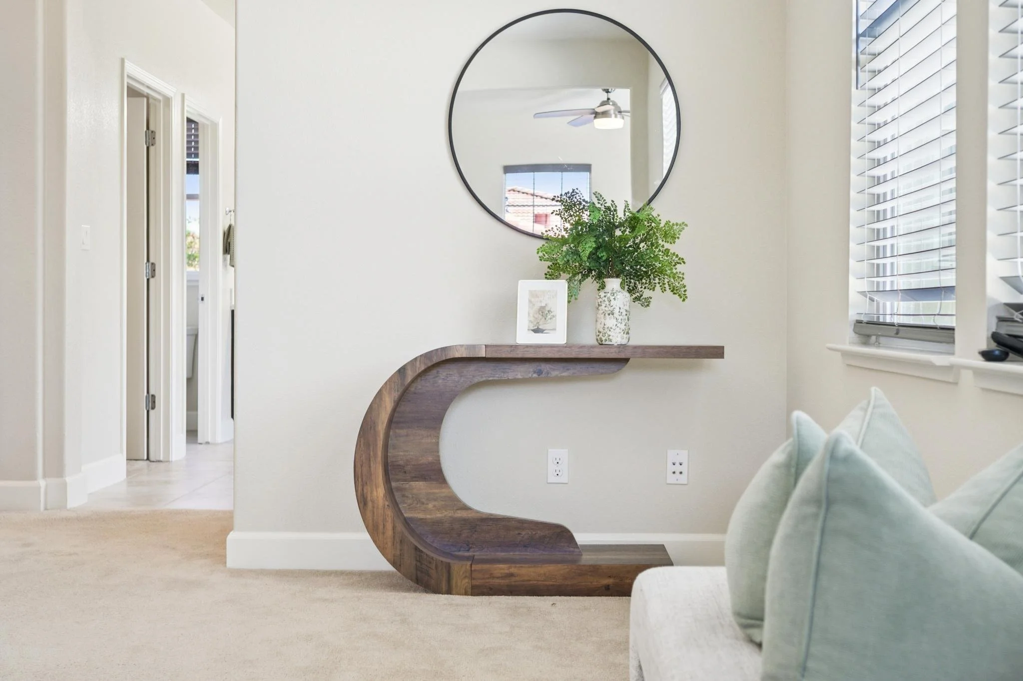 Interior view of a home living space with a decorative wooden console table, a round mirror, a potted green plant, and a picture frame on the table. A partial view of a light-colored sofa with pillows is visible in the foreground, and a window with b
