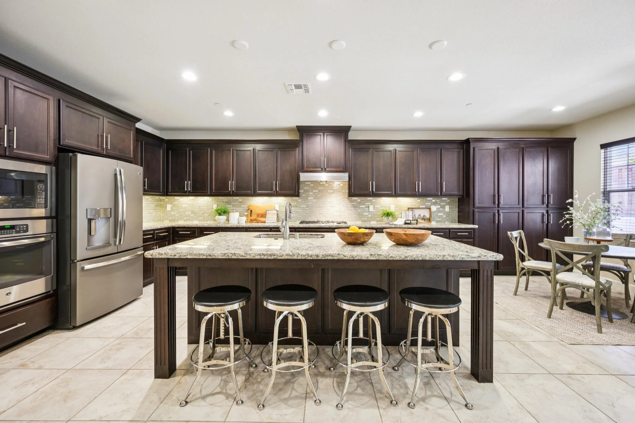 Modern kitchen with dark wooden cabinets, granite island, stainless steel appliances, tiled backsplash, dining table with chairs, and potted plants near a window.