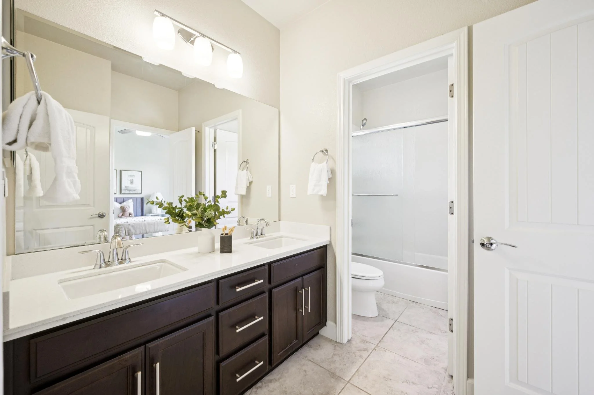 Bathroom with dual sinks, large mirror, dark wooden cabinets, and a walk-in shower with white door. There are white towels on hooks and a plant on the counter.