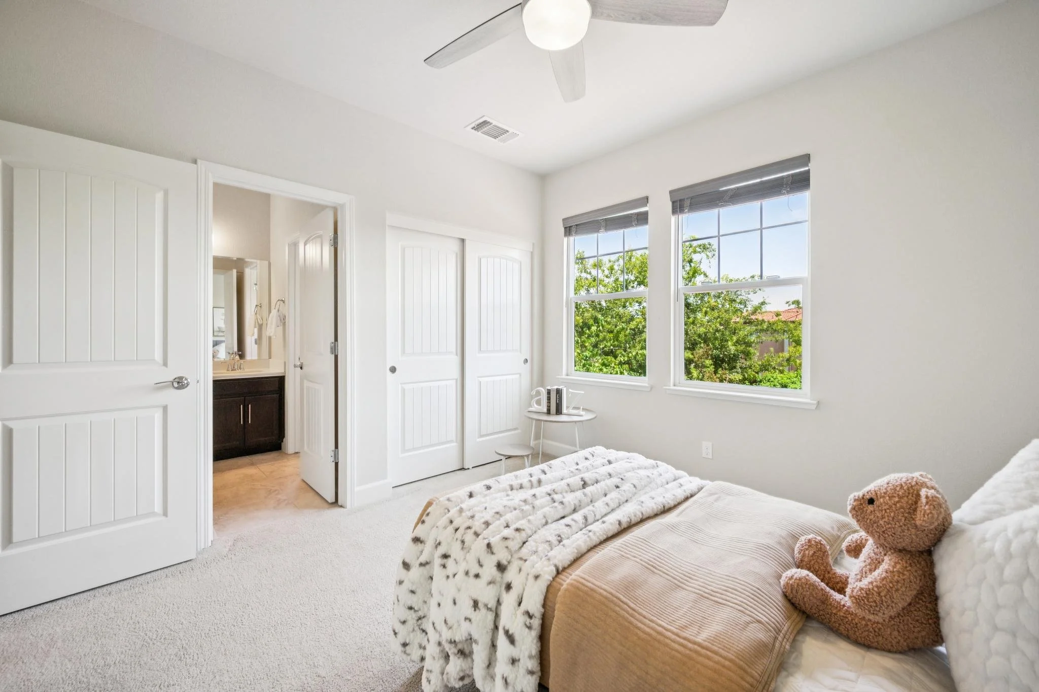 A bedroom with two windows showing trees outside, a bed with a plush teddy bear, a throw blanket, and a small side table with decorative items. There is a ceiling fan with a light and two closed closet doors.