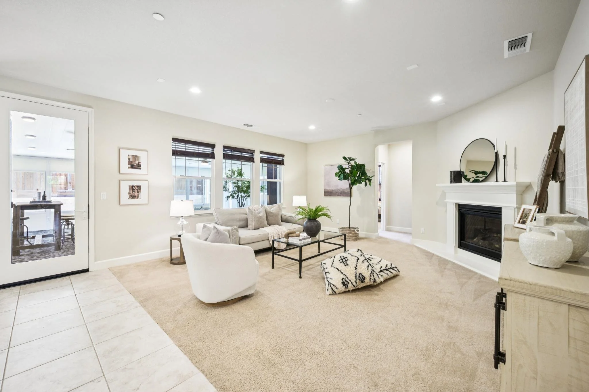Living room with beige carpet, white walls, a beige sofa with throw pillows, a glass coffee table, a white armchair, a tall potted plant, and a fireplace with a round mirror above it.