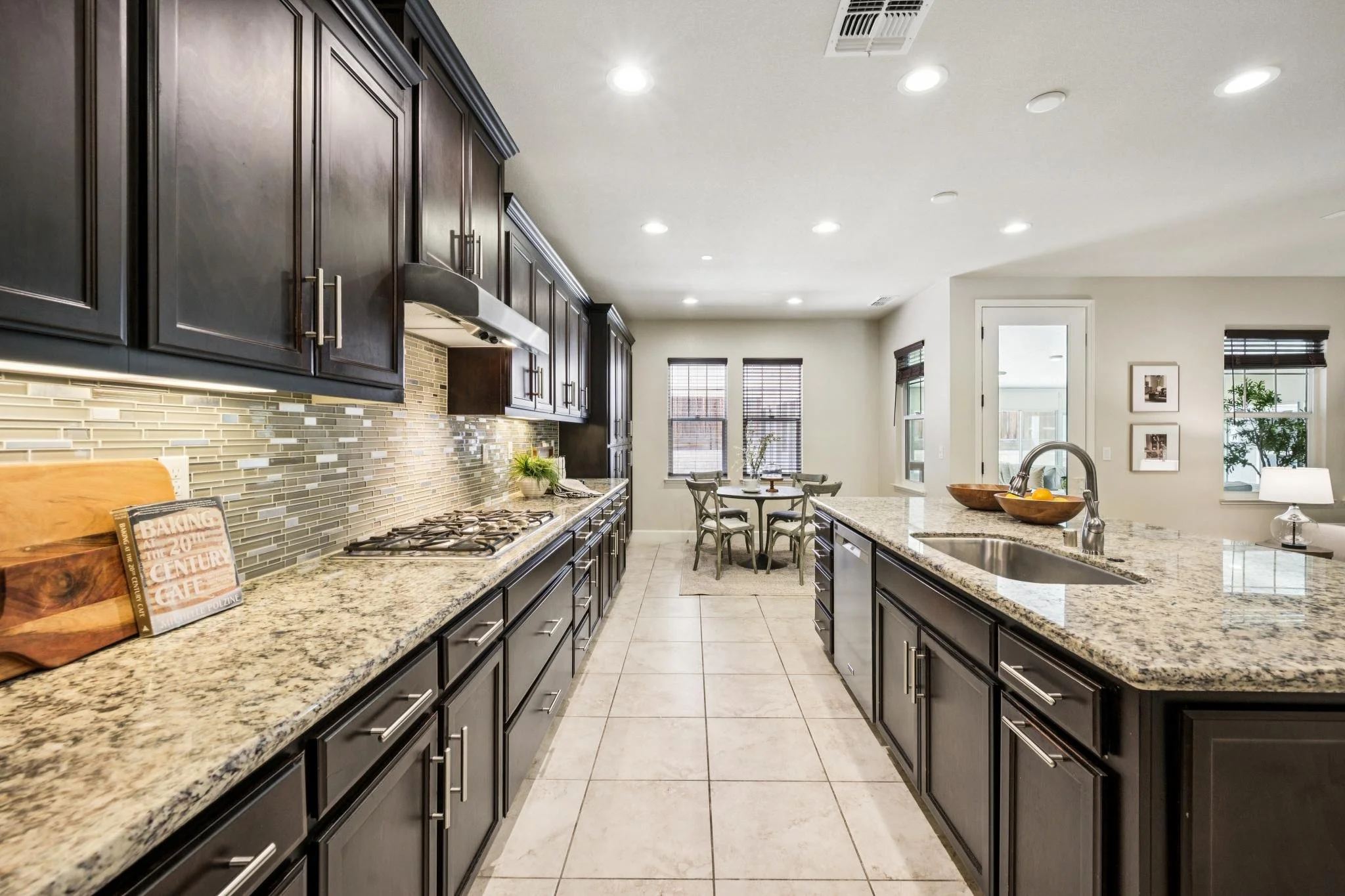 Bright kitchen with dark wooden cabinets, granite countertops, tile backsplash, and stainless steel appliances, including a stove and a sink. A small dining area with four chairs is visible in the background, along with large windows and some wall de