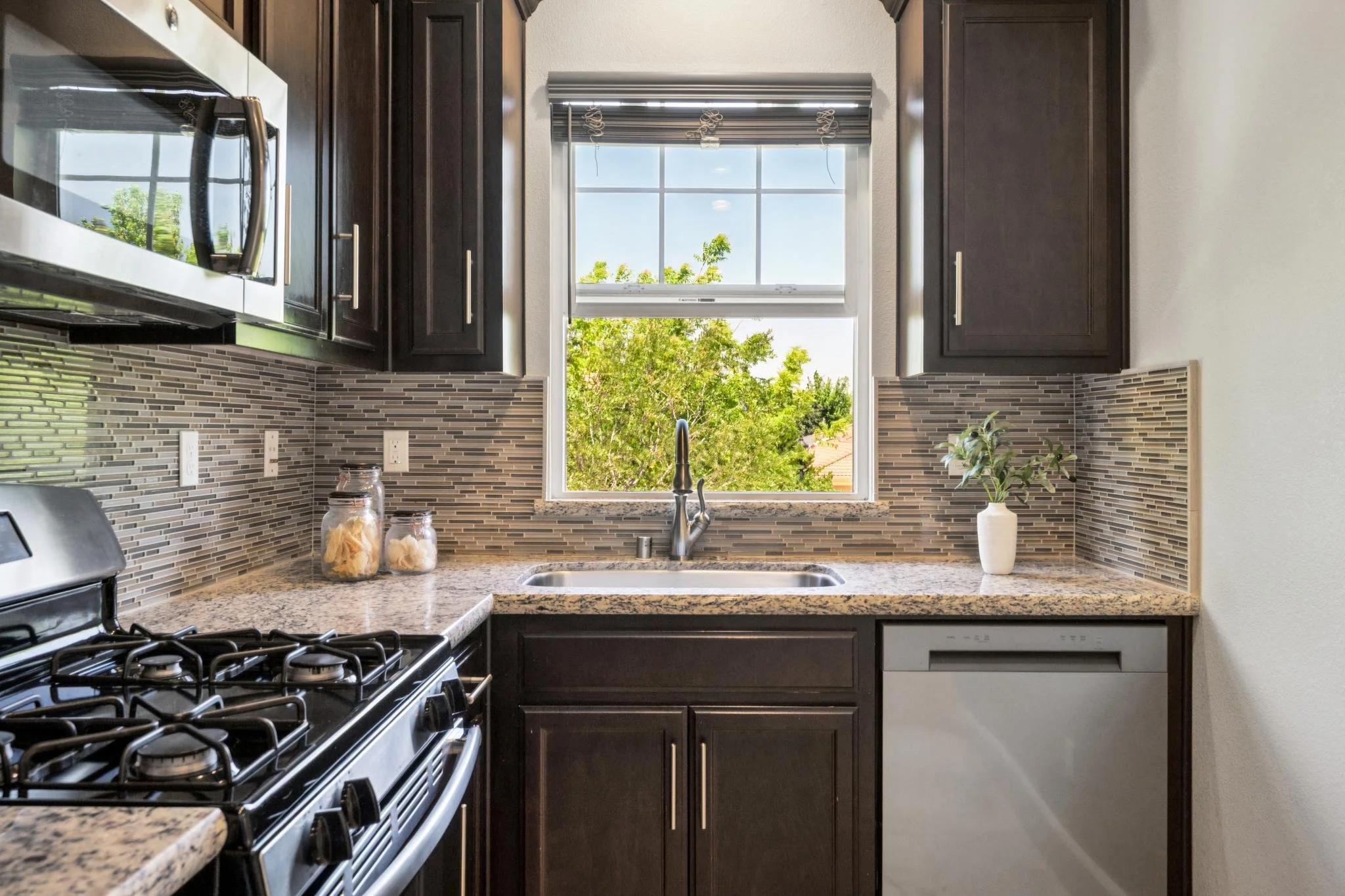 Kitchen with dark wood cabinets, granite countertops, a window with trees outside, and a stove with jars on the counter.