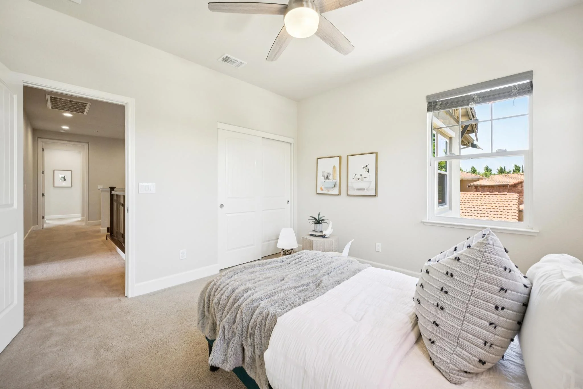 A bright bedroom with a large window showing rooftops outside. The room has white walls, hanging framed artwork, and a bed with pillows and a textured blanket. A ceiling fan with a light fixture is mounted on the ceiling.