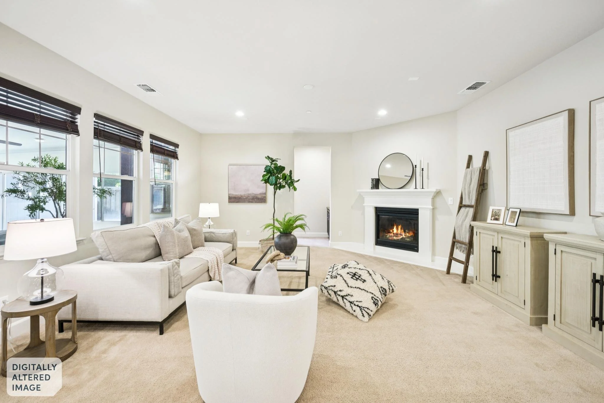 A bright living room with white walls, large windows with black blinds, a fireplace with a fire, a beige sofa with cushions, a white armchair, a coffee table, a potted plant, framed artwork, and decorative items.