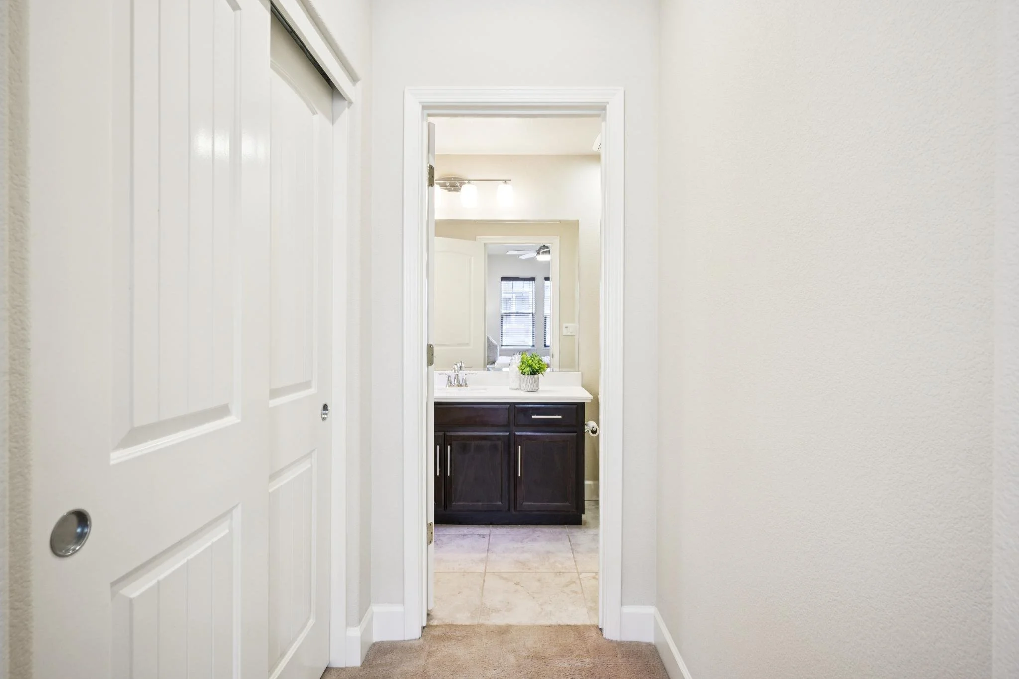 View through a doorway into a bathroom with dark wood vanity, white countertop, and a plant. Bright room with window and ceiling fan.