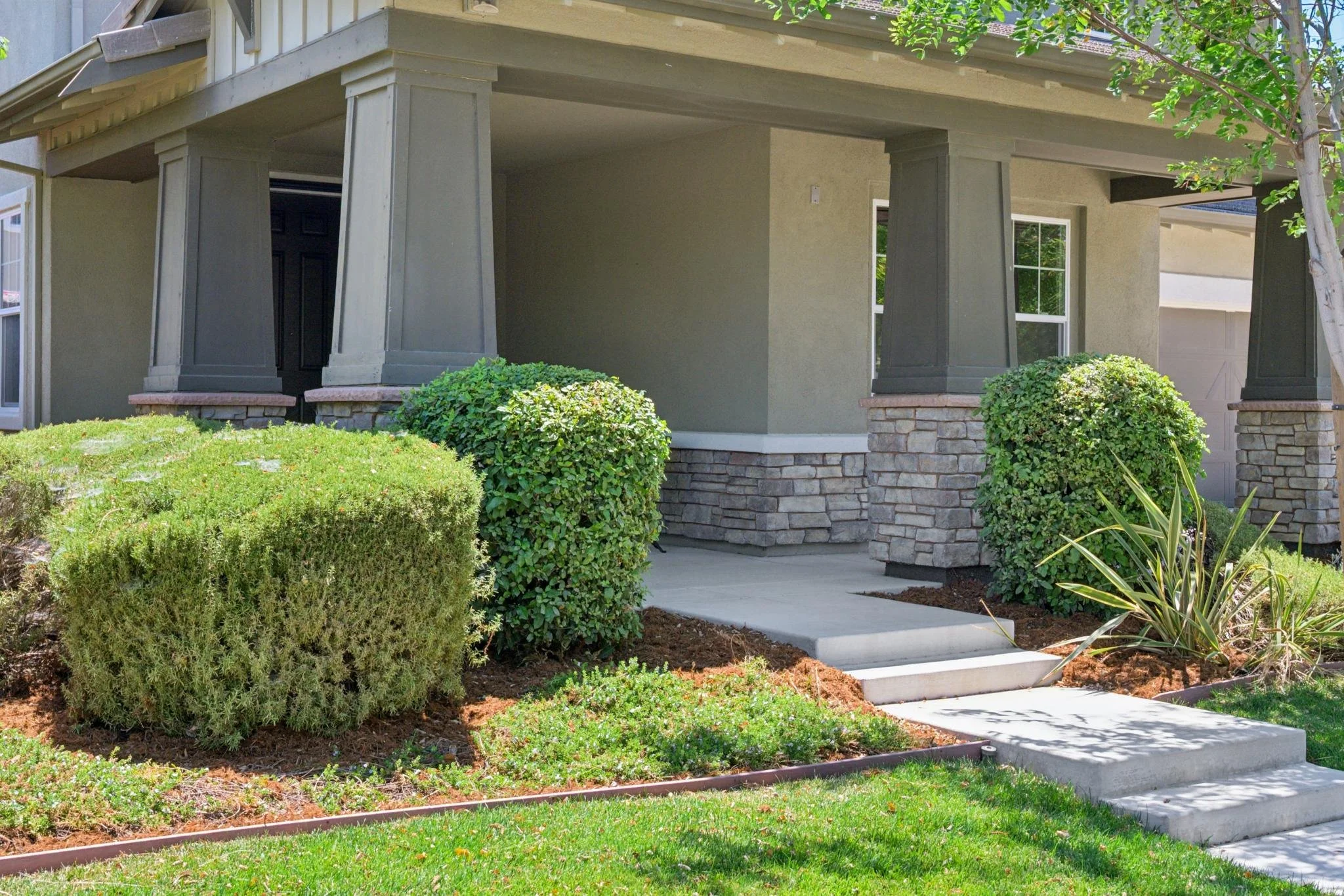 Front porch of a house with a concrete walkway, steps, and bushes in the yard. The house features a covered porch with stone-paved columns and a beige exterior wall with white window frames and dark shutters.