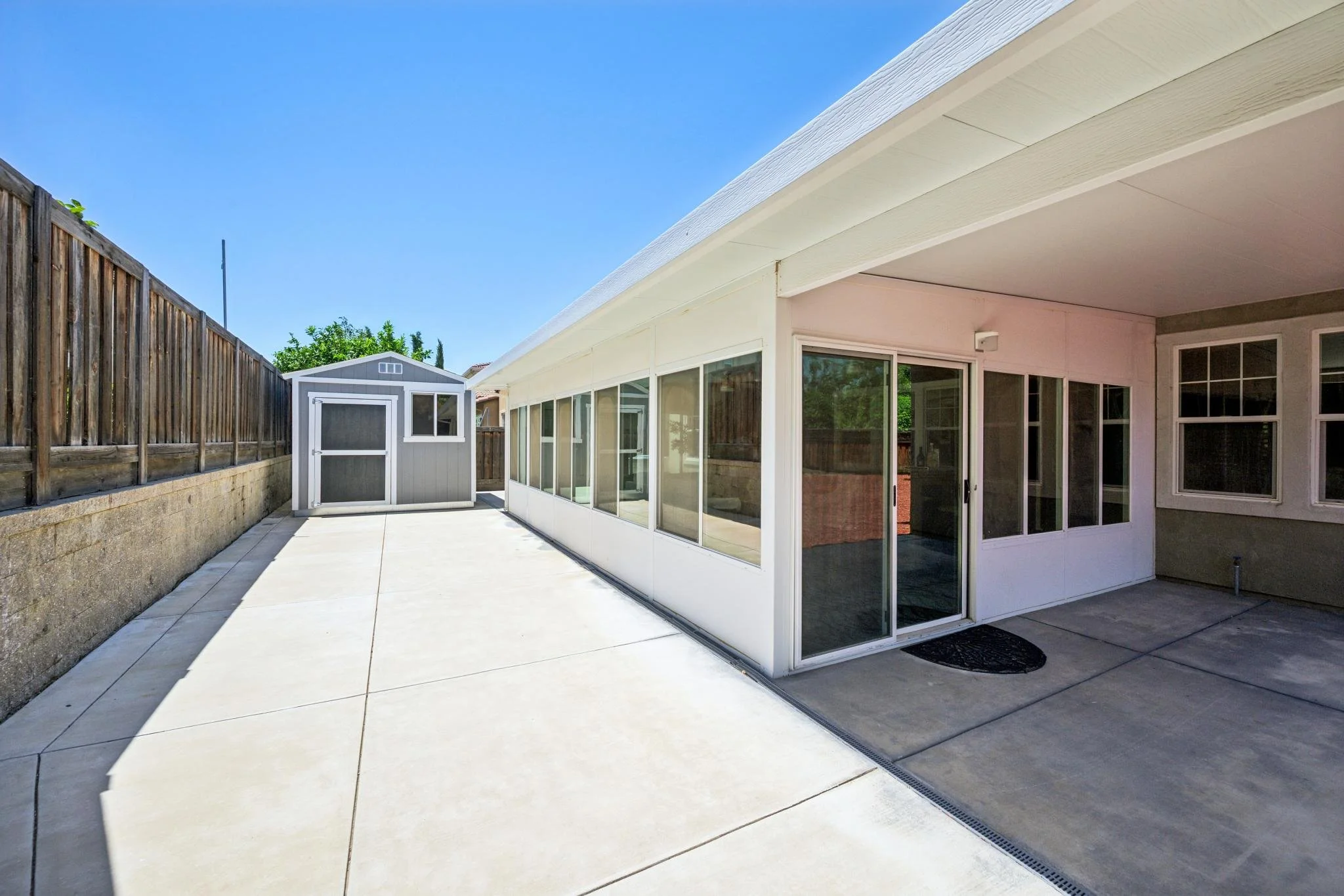 Backyard patio with a concrete floor, a white screened-in porch, a small shed, a wooden fence on the left, and clear blue sky overhead.