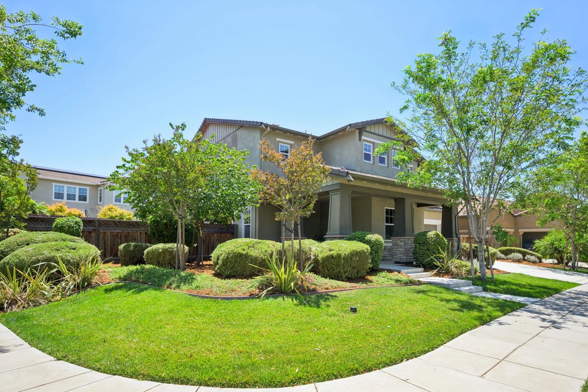 A neatly landscaped front yard with green grass, small bushes, and trees in front of a beige two-story house with a gray roof and visible windows, under a clear blue sky.