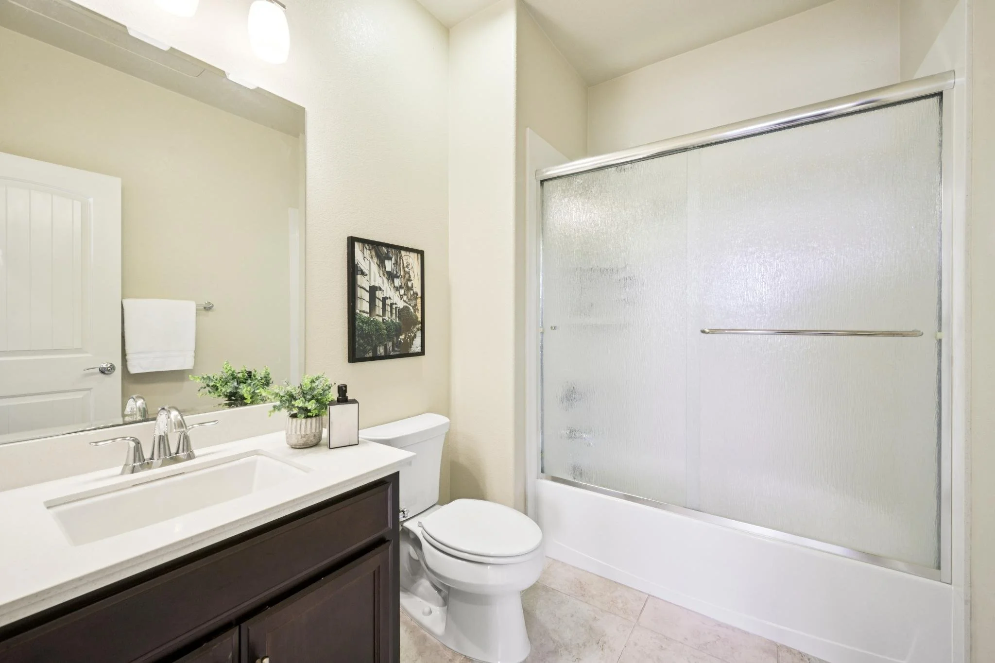 A bathroom with a vanity mirror, a plant on the white sink counter, a framed picture on the wall, a toilet, and a frosted glass shower door.