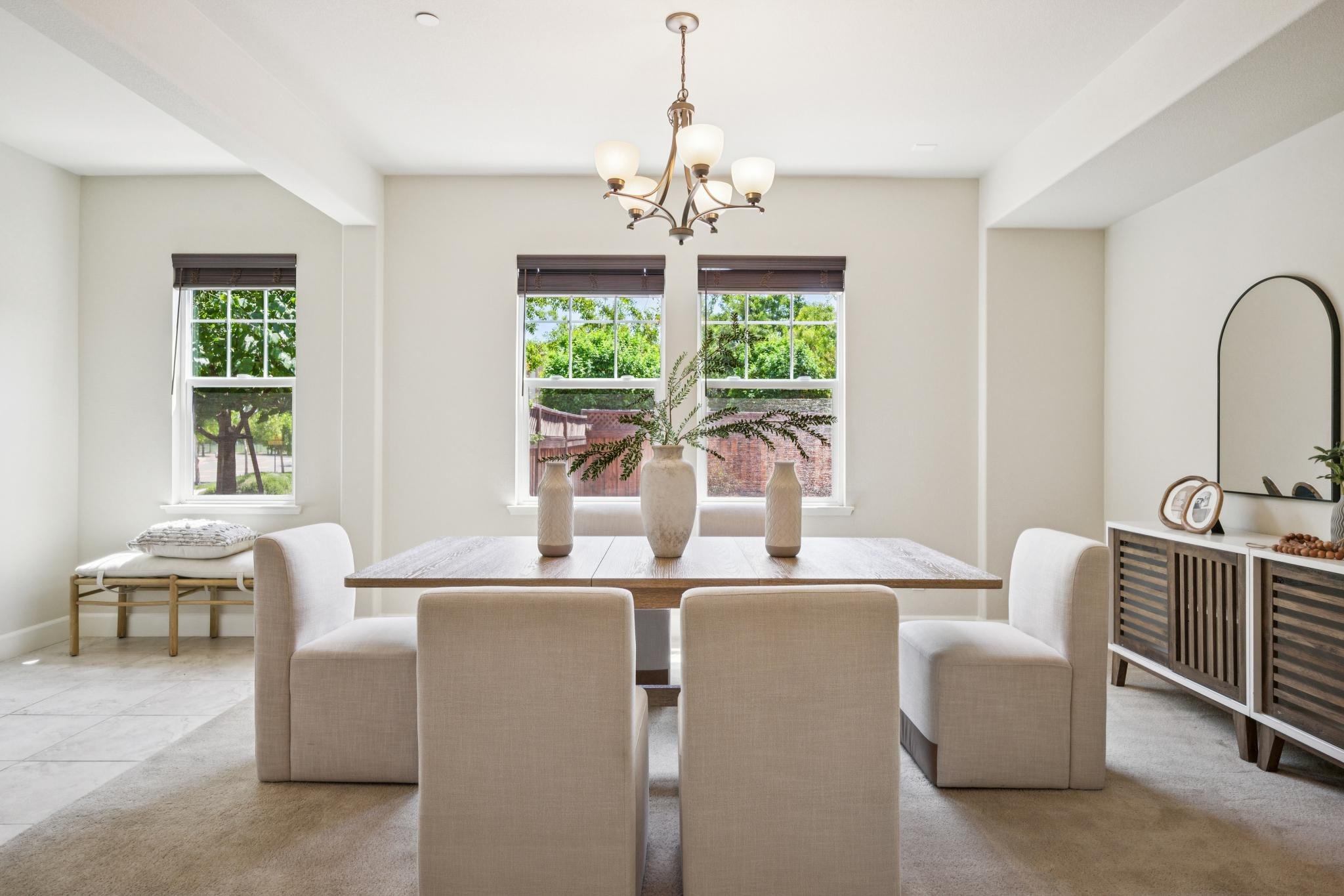 Bright dining room with large window, white chairs around a wooden table decorated with three white vases, chandelier above, and window blinds.