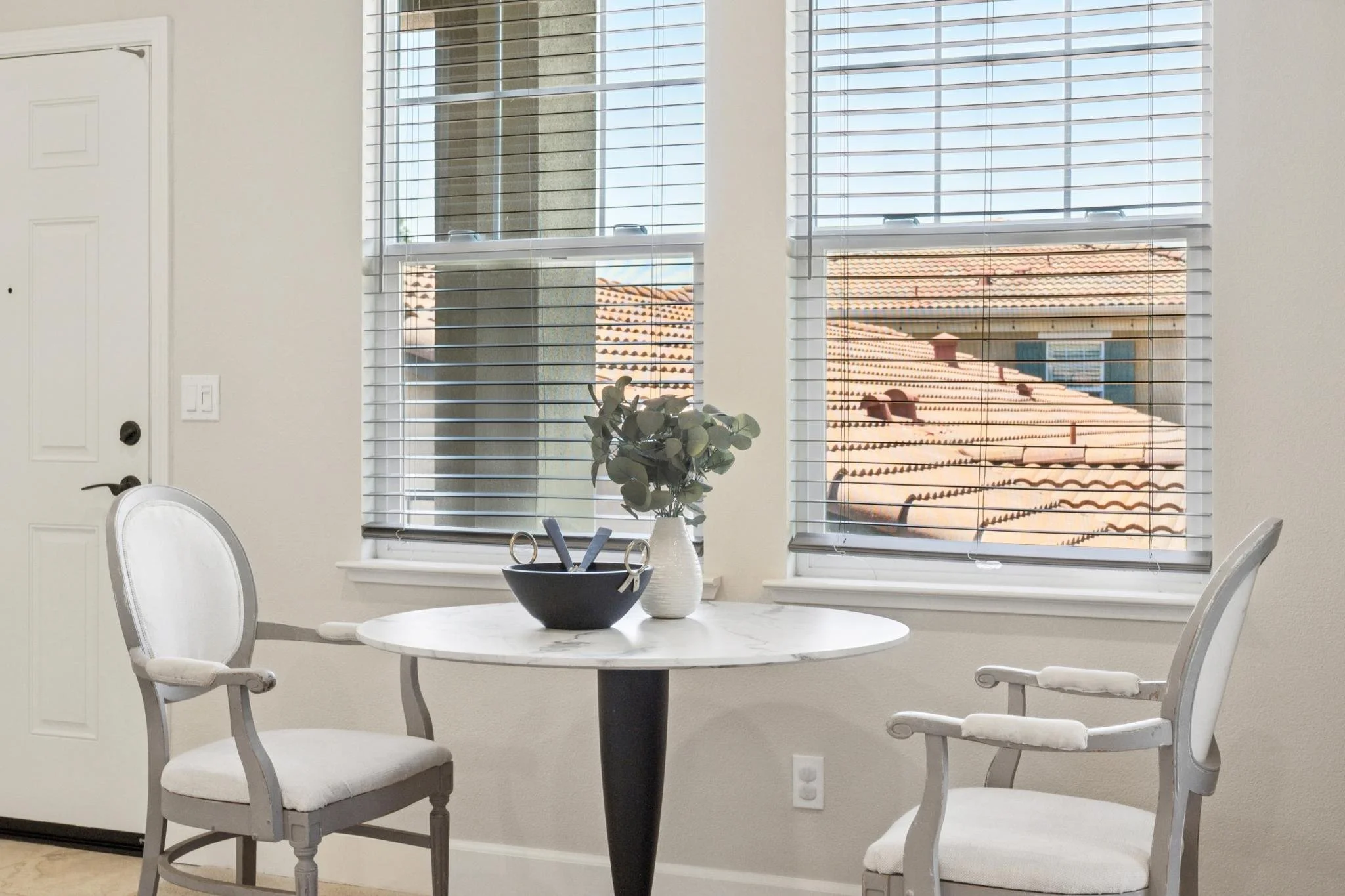 A dining room with a white round marble table, three white chairs with padded seats, and a window with white blinds showing rooftops and blue sky outside.