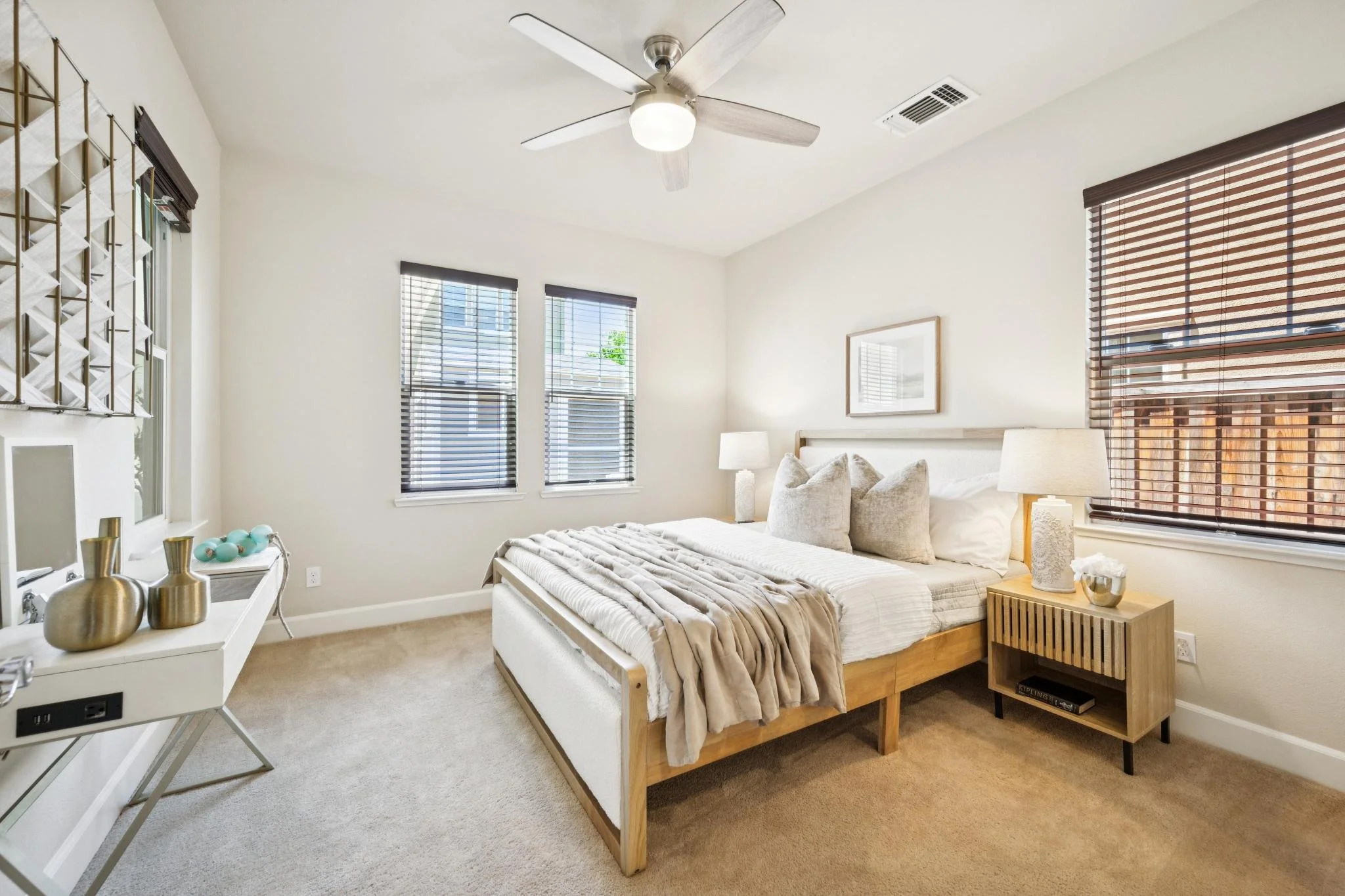 A cozy bedroom with a white bed, beige bedding, and multiple pillows. There are three windows with blinds, and a ceiling fan with lights. A nightstand with a lamp and decorative items is beside the bed. There's a white console table with vases and de