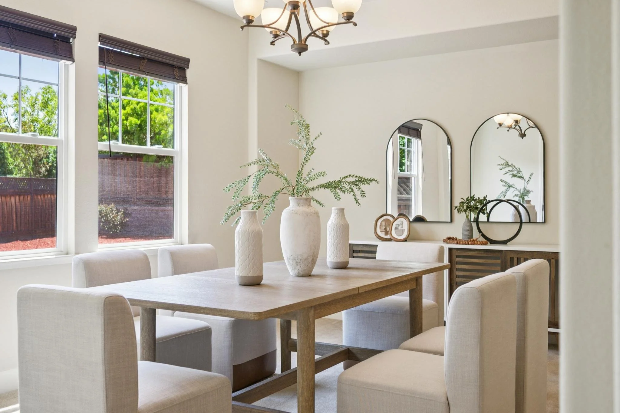 A dining room with a wooden table, surrounded by six white upholstered chairs, decorated with three white vases holding greenery, and a wall mirror, with windows and a chandelier overhead.