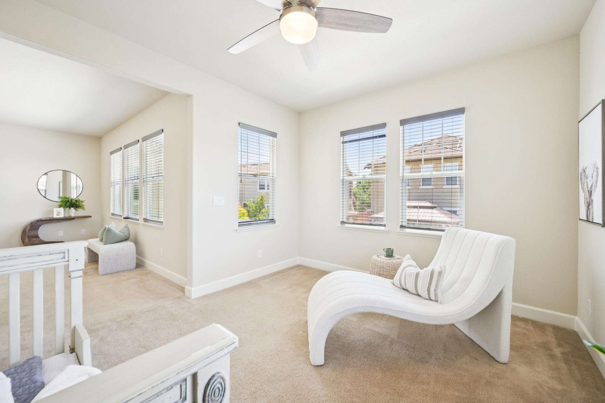 Living room with beige carpet, white walls, multiple windows with blinds, a ceiling fan with light, white curved lounge chair, small wicker side table with a green cup, and wall art featuring an animal illustration.