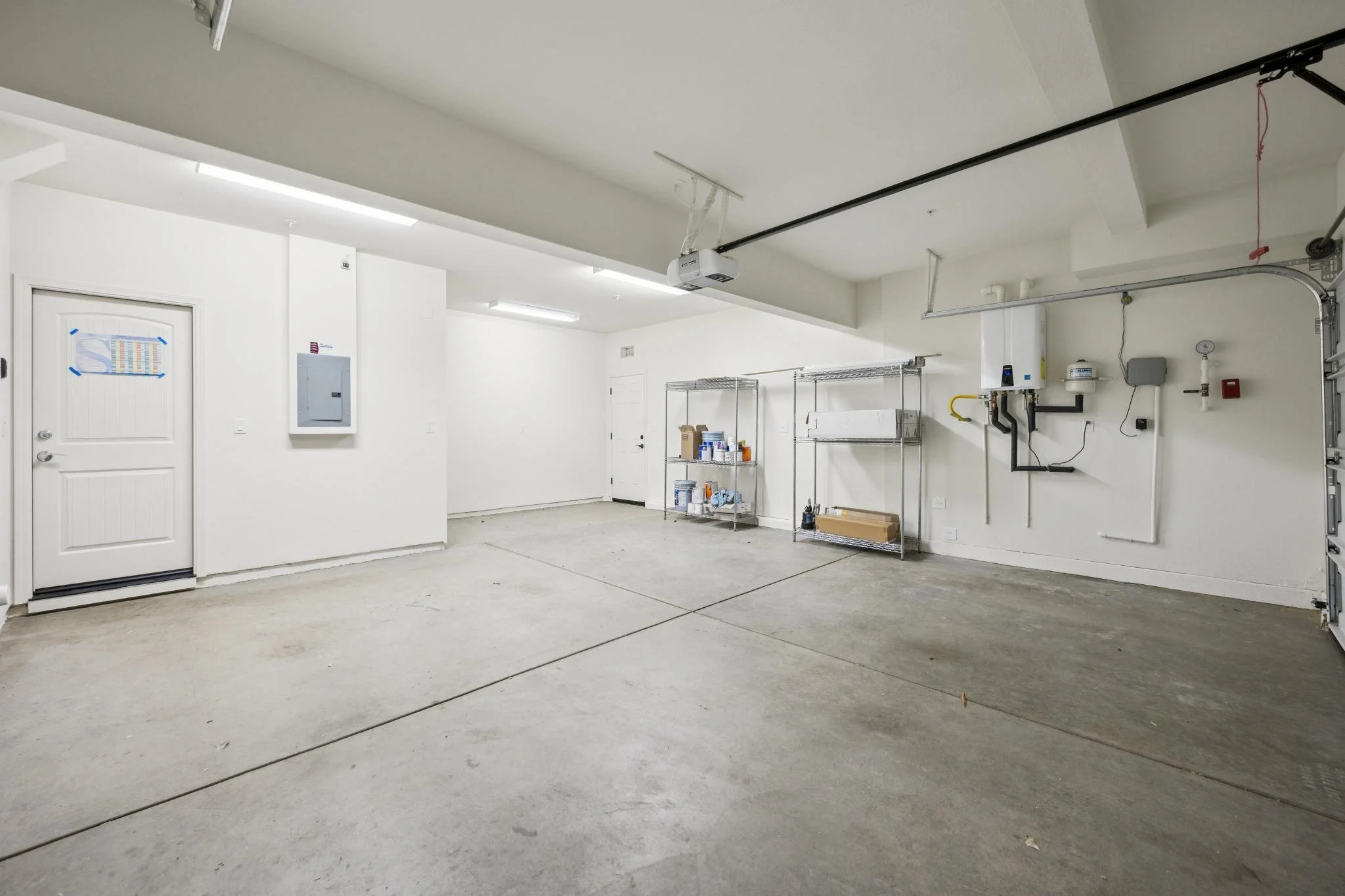 Empty garage with concrete floor, white walls, ceiling-mounted garage door opener, shelves with paint and supplies, and electrical panels.