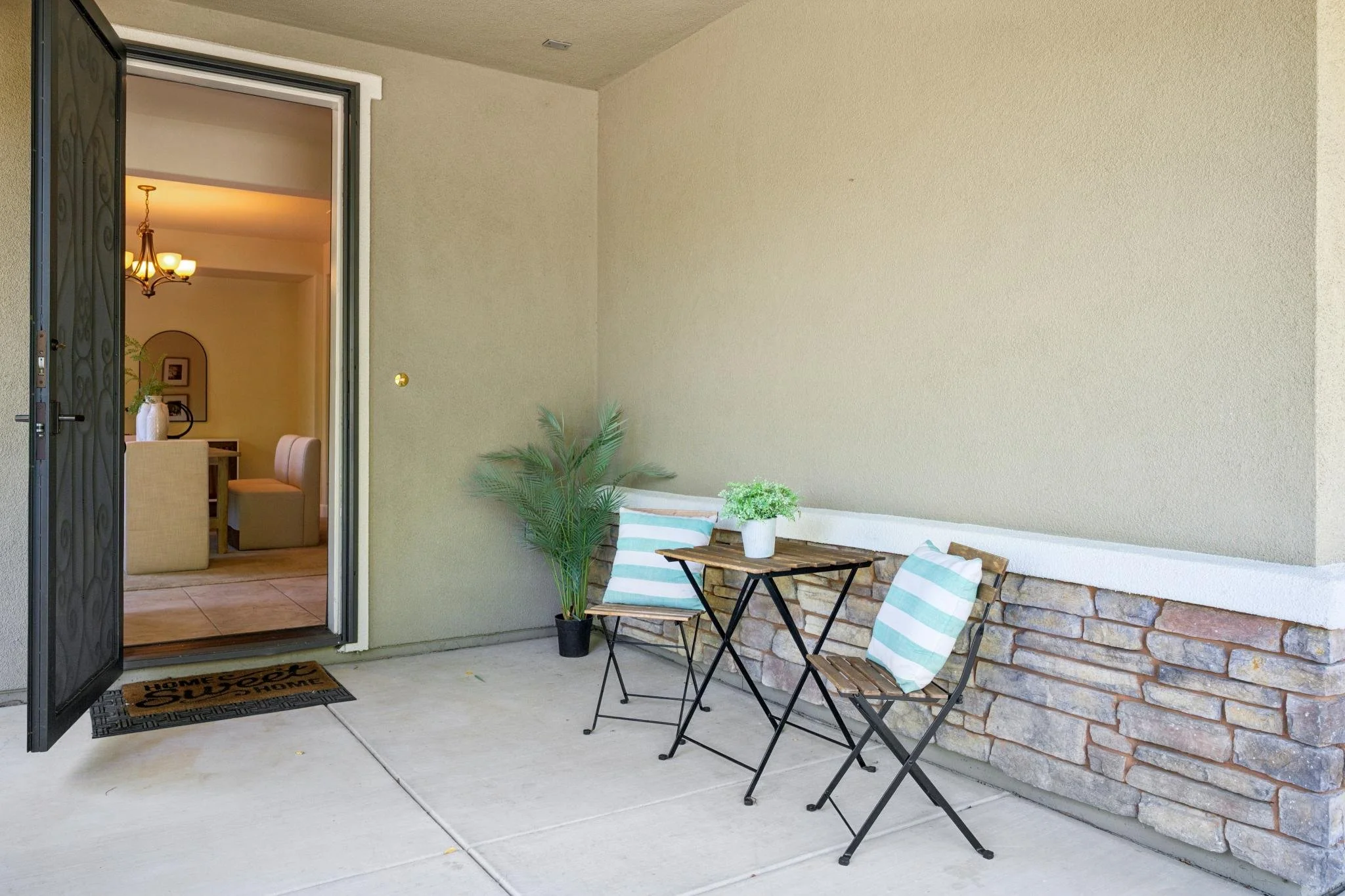 Front porch with two chairs, a small table with plants, and an open screen door leading into a dining area with a chandelier.