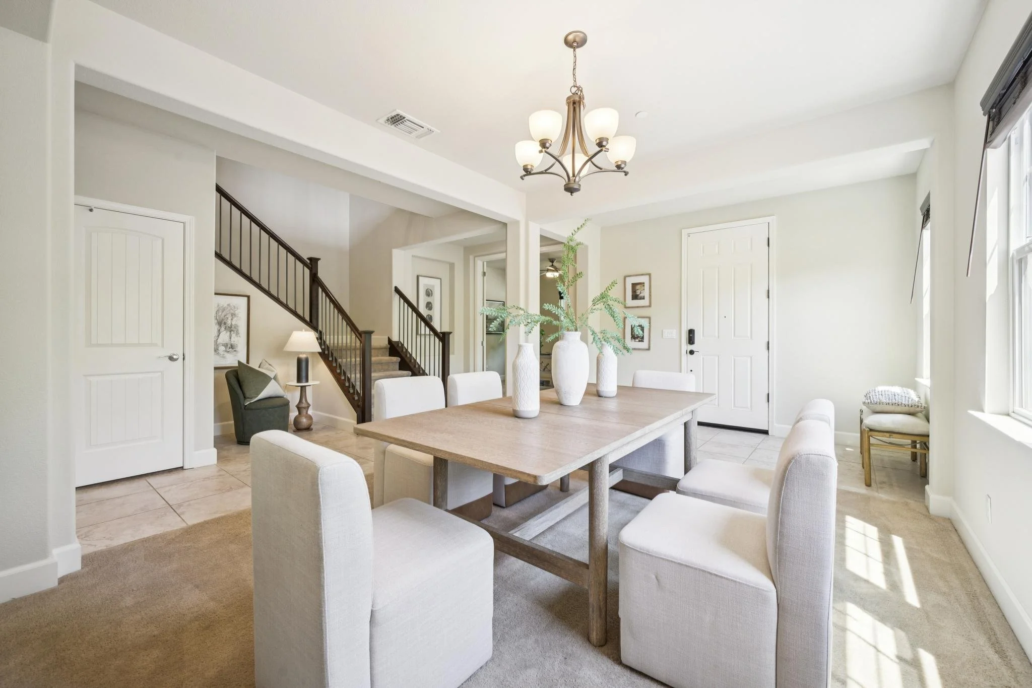 Bright dining area with a wooden table, white upholstered chairs, and multiple tall white vases with green plants. A staircase leading upstairs is visible on the left, and there's a window on the right with sunlight streaming in. The ceiling has a ch